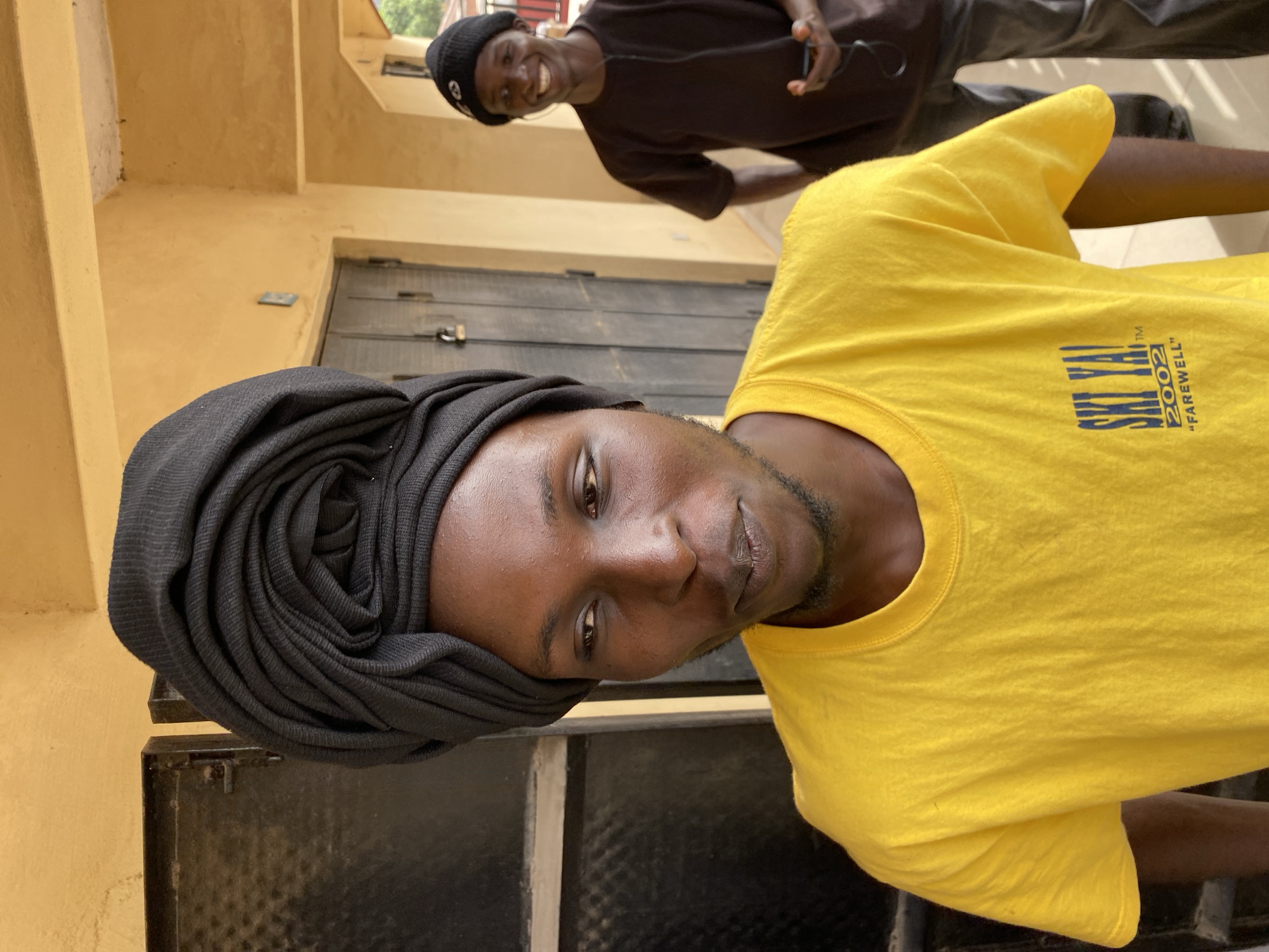 The image depicts two young men outdoors in what appears to be Jos, Nigeria. One man is in the foreground, wearing a yellow t-shirt with the words "SKI VAL 2002 'Farewell'" printed on it. He has a black cloth wrapped around his head and is partially in front of what appears to be a black metal structure or door. The second man is in the background, wearing a black beanie and a brown shirt, holding what looks like a phone or music player. They are standing outside a building with painted yellowish walls. The overall scene suggests a casual setting.
