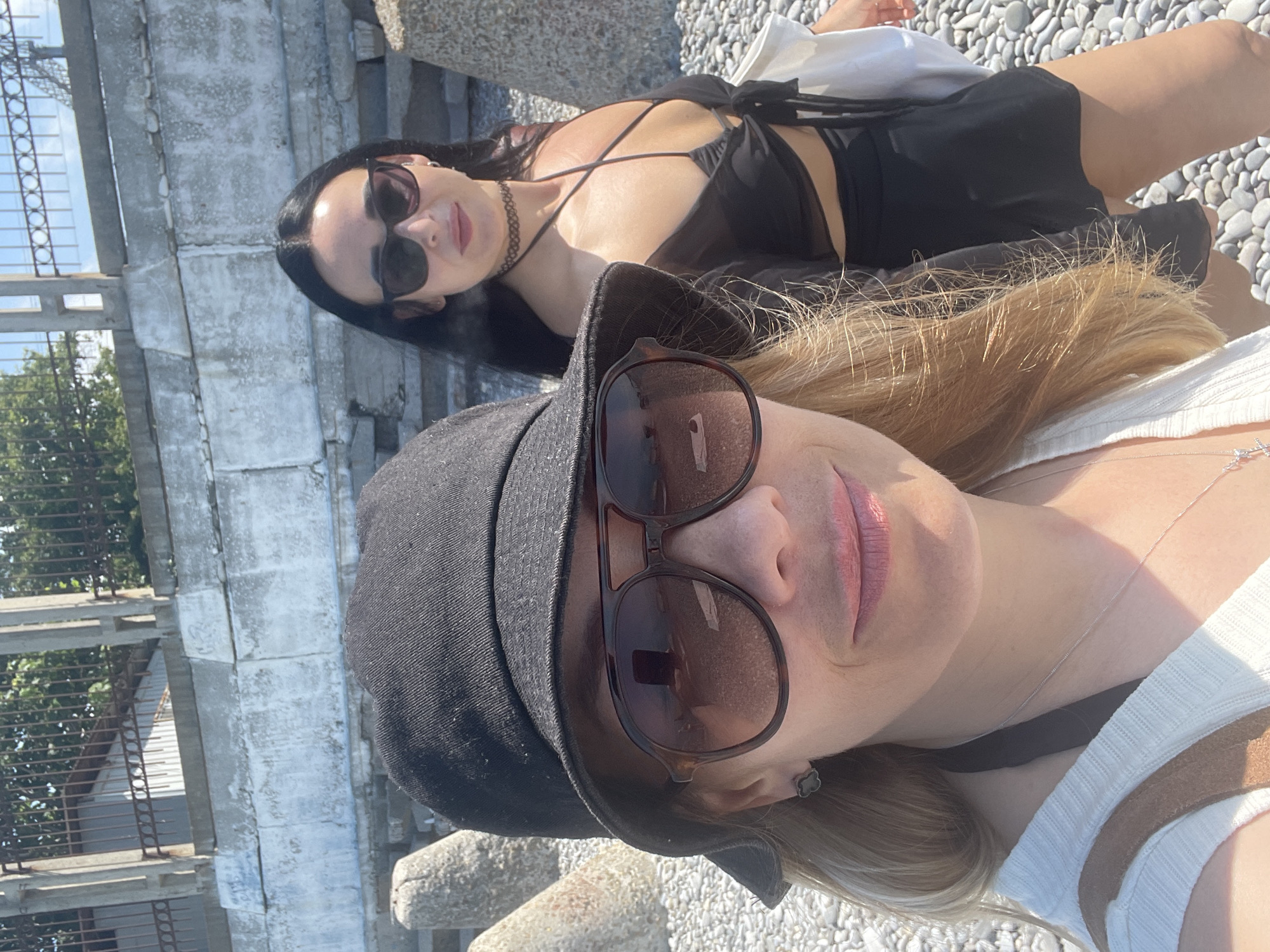 Two women are standing on a pebble beach near a concrete wall and metal fence. The woman in the foreground is wearing a black bucket hat and large sunglasses, with blonde hair visible. She is wearing a white top with brown straps and a necklace. The woman in the background is wearing sunglasses, a black choker, a black bikini top with a sheer black wrap tied at the front, and a black skirt. She has dark hair. Both women are looking towards the camera. There are concrete barriers on the beach. The background shows a sunny day with some greenery behind the fence.