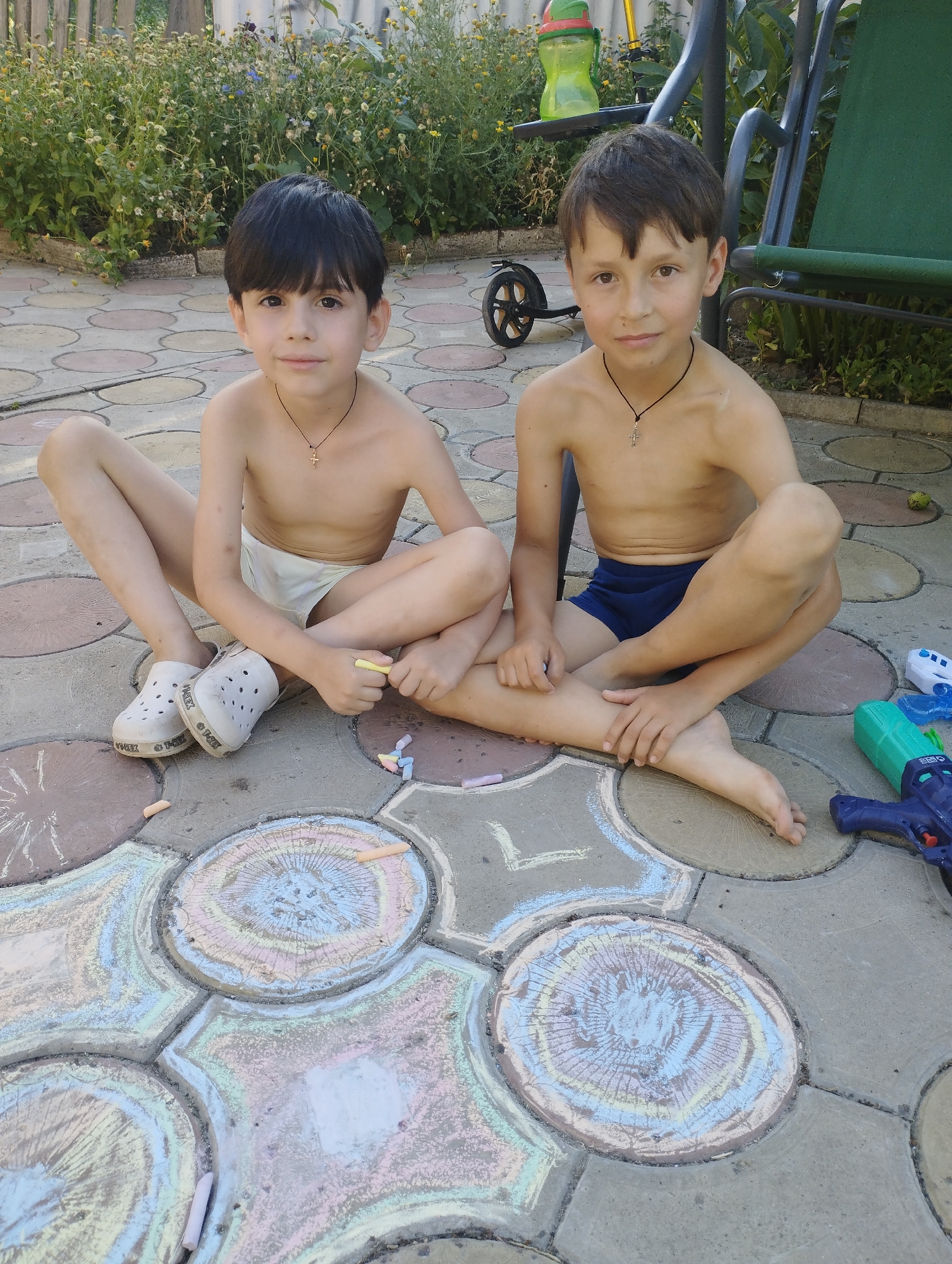 Two young boys are sitting on a paved patio, drawing with chalk. The boy on the left has dark hair and is wearing white shorts and white clogs. He is holding a yellow piece of chalk and is focused on the drawing in front of him. The boy on the right has brown hair and is wearing blue swim trunks. He is holding a piece of chalk in his left hand and is looking towards the camera with a slight smile.
In front of the boys, the patio pavers have been decorated with colorful chalk drawings. The drawings appear to be abstract, with swirling patterns and lines in various shades of pink, blue, yellow, and white. There are also several pieces of chalk scattered on the ground.
Behind the boys, there is a garden area with lush green plants and some small yellow and purple flowers. A green swing set is visible in the background, along with a green water bottle on a shelf attached to it. A black wheel is also visible near the swing set.
The scene appears to be set in a backyard on a sunny day. The boys are engaged in a creative activity, and their expressions suggest they are enjoying themselves. The overall impression is one of a casual and pleasant summer afternoon. There are no visible text elements in the image.