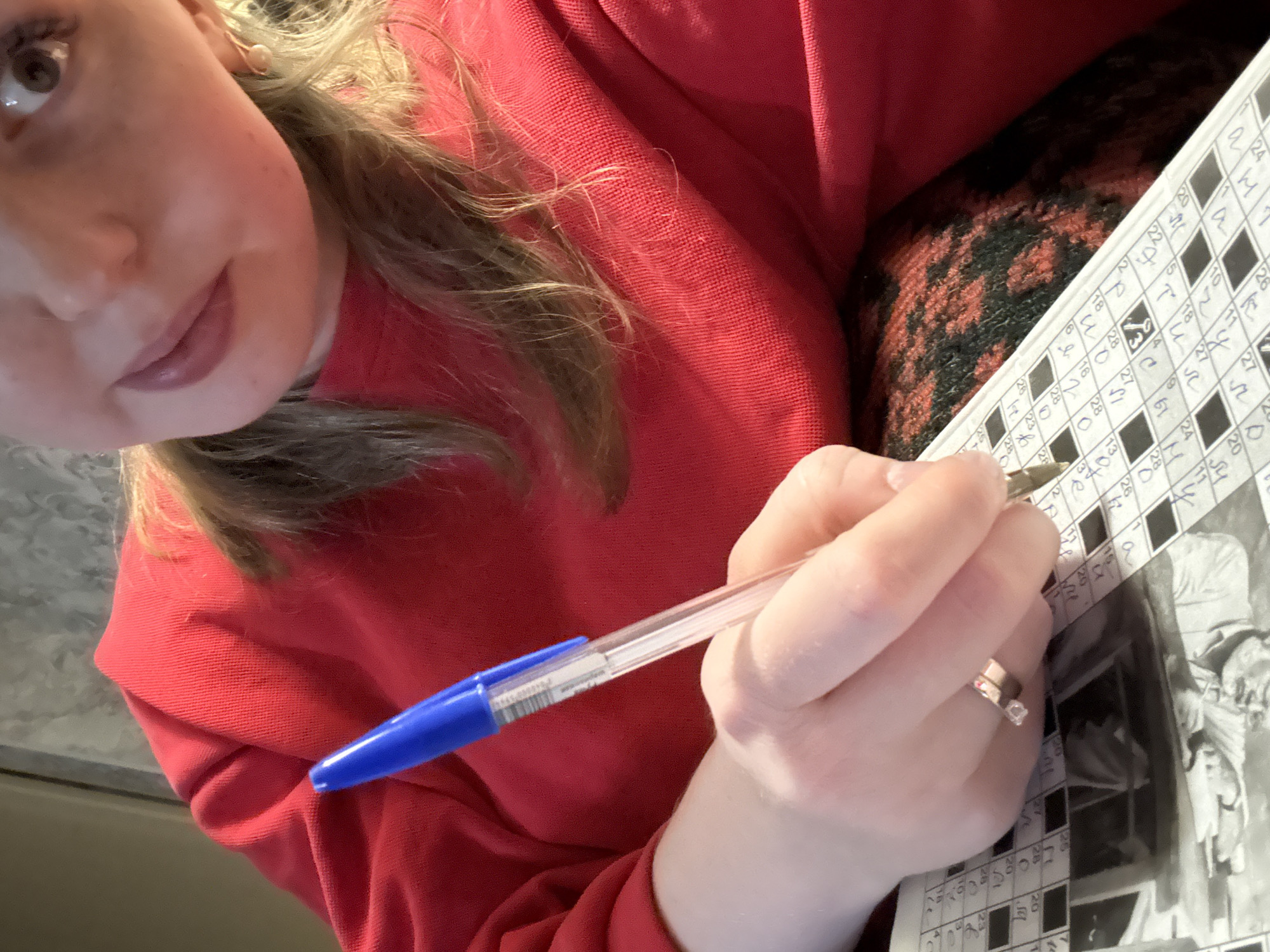 An overhead, slightly tilted view captures a person engrossed in solving a crossword puzzle. The primary subject is an individual, likely female given the facial features and visible jewelry, seen from the nose down, with her right hand holding a blue ballpoint pen. She is wearing a red, possibly knitted or ribbed, long-sleeved garment, perhaps a sweater or hoodie. Her long hair, a mix of light and darker brown shades, is visible around her face and shoulder. A subtle pearl earring is discernible in her left ear, and a ring with a clear stone adorns her right ring finger.
The pen, a transparent barrel with a blue cap and tip, is positioned over a white crossword puzzle page. The page features a grid with black and white squares, some filled with numbers and letters. Visible numbers in the grid include "4", "20", "22", "18", "3", "28", "27", "61", "24", "20", "26", "23", and "191280". The letter "Q" is visible in one square. Below the crossword grid, a monochrome image, possibly an old photograph, is partially visible on the page.
The person appears focused, with her eye looking towards the puzzle. Part of a patterned fabric, perhaps a cushion or blanket with black and reddish-brown hues, is visible beneath her arm and the puzzle book. The background is mostly blurred, offering minimal location cues beyond indicating an indoor setting.