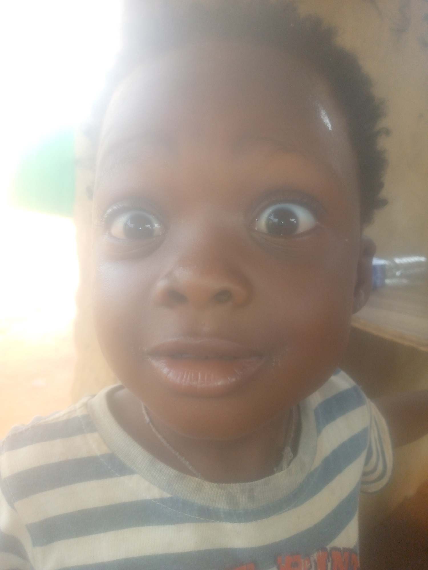 A close-up, overexposed portrait features a young child, likely a toddler or preschooler, looking directly into the camera. The child has dark skin, wide-open, dark brown eyes expressing surprise or curiosity, and short, dark, curly hair. Their mouth is slightly open, revealing full lips. A faint white reflection or fleck is visible on their forehead.
The child is wearing a light-colored t-shirt with horizontal blue and white stripes. A thin, silver-colored necklace or chain is visible around their neck. The lower part of the shirt also shows some blurry, indecipherable red text.
The background is heavily overexposed, especially on the left side, where a bright, washed-out light source dominates. A blurry, green shape is faintly discernible within this bright area. On the right, the background is darker and indistinct, suggesting an indoor or sheltered setting with some wooden elements, possibly furniture or a wall. The overall lighting is very bright, contributing to the hazy, ethereal quality of the image. The photo captures a moment of wide-eyed wonder or startled attention from the child.