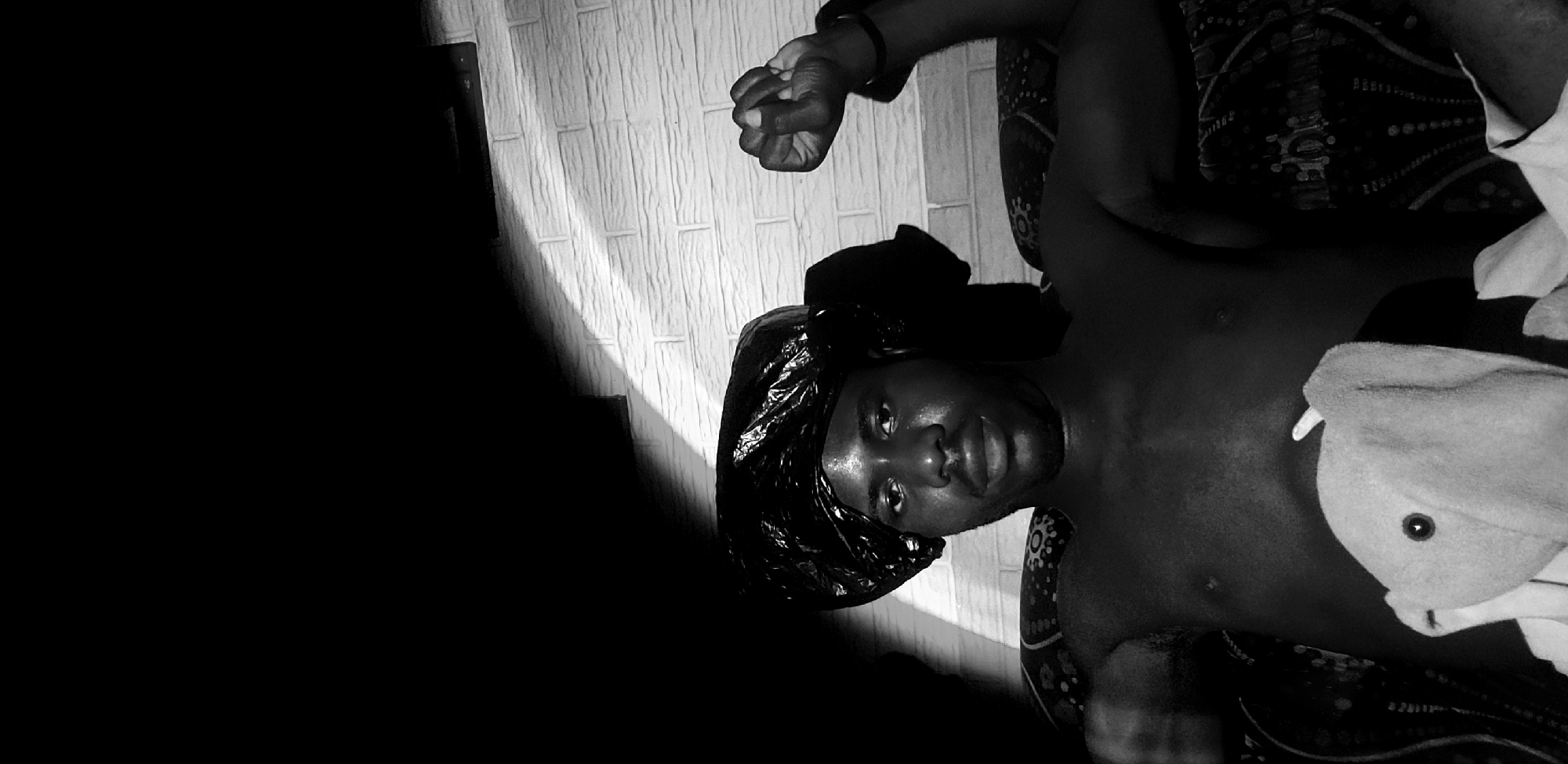 In Narayi, Nigeria, a shirtless man with a dark head covering sits on a patterned couch. The lighting casts dramatic shadows across his face and the brick-patterned wall behind him. His right arm is raised, with his fist clenched. A light-colored towel is draped over his lap. The overall scene is stark and monochromatic.