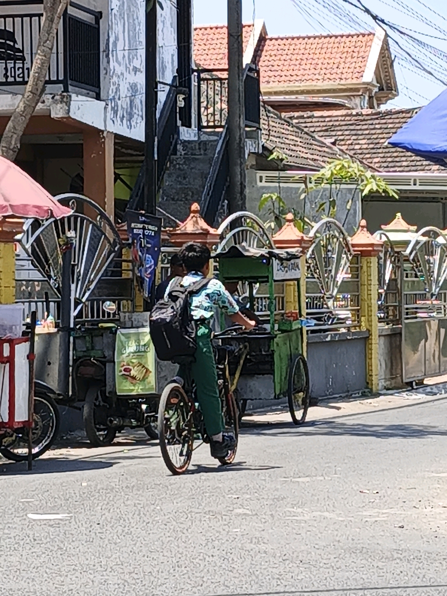 In Ketegan, Indonesia, a young boy wearing a green and white patterned shirt and dark green pants rides a bicycle with a backpack. He is positioned in the middle of a paved road, and a food cart is attached to the back of his bicycle. The food cart is green and has a yellow sign on it. To the left of the boy, another food cart with a red and white striped awning is visible. Behind the food carts, there is a building with a grey facade and decorative metalwork. Further in the background, a house with a red tile roof and an outdoor staircase can be seen. The sky is clear and blue.