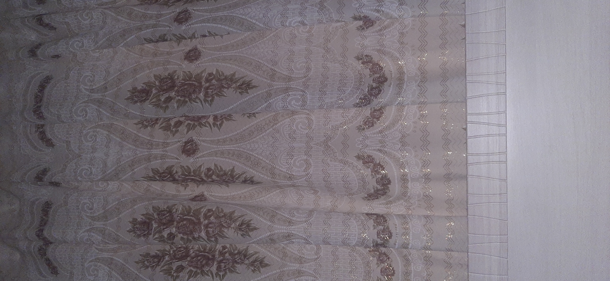 The image shows a close-up view of a beige curtain with a patterned design. The pattern features vertical floral motifs with roses and leaves in a muted brown or grey color. Interspersed between the floral elements are paisley-like shapes and decorative borders with small, shimmering accents that appear to be glitter or metallic thread. The fabric has a textured appearance, with some areas of sheer material and others appearing more opaque. The bottom portion of the image shows a band of fabric with vertical pleats or stitching, followed by a solid, lighter colored section, possibly a different fabric or the wall behind the curtain. The overall impression is of a decorative textile, likely an interior furnishing. There is no indication of a city, location, or any specific context beyond the object itself.