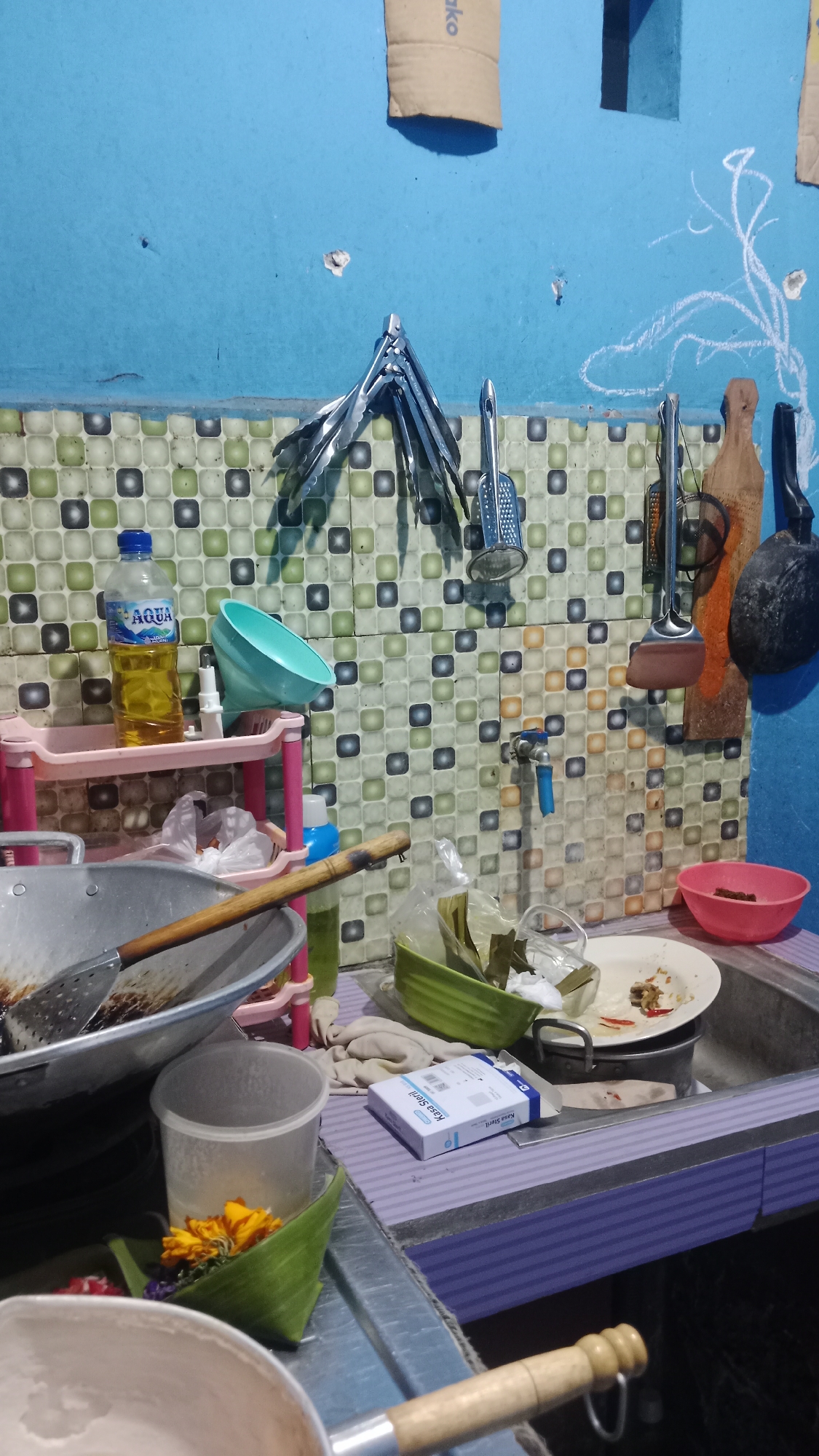The image depicts a cluttered kitchen counter in Denpasar, Indonesia. The counter is filled with various kitchen utensils and food items. In the foreground, a wok is visible, along with a strainer and a wooden-handled utensil. To the right, a sink is partially filled with dirty dishes. A faucet is mounted on a tiled backsplash, which is adorned with a mosaic of square tiles in various shades of green, beige, and orange. Several cooking tools, including tongs and spatulas, are hanging on the wall above the sink. A bottle of Aqua brand cooking oil stands on a pink shelf unit, next to a blue funnel. A packet labeled "Kasa Steril" (Sterile Gauze) is placed on the counter near the sink. The walls are painted blue, and some areas show signs of wear, with small holes and chalk drawings. The overall impression is of a functional but untidy kitchen space.
