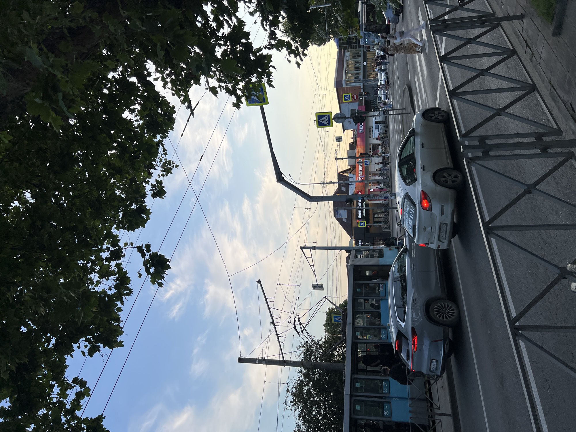 In Krasnodar, Russia, a wide street features a light blue tram with passengers boarding, moving from left to right. In front of the tram, a silver SUV is parked on the road. Further ahead, a white sedan is visible, with its brake lights illuminated, in motion. The street is lined with shops and buildings, including a "Magnit" store. Two women are crossing the street at a pedestrian crossing. Overhead, a network of tram wires is strung between poles and across the street. Trees with green leaves overhang the scene from the top of the frame. The sky is a pale blue with scattered clouds, indicating either early morning or late afternoon.