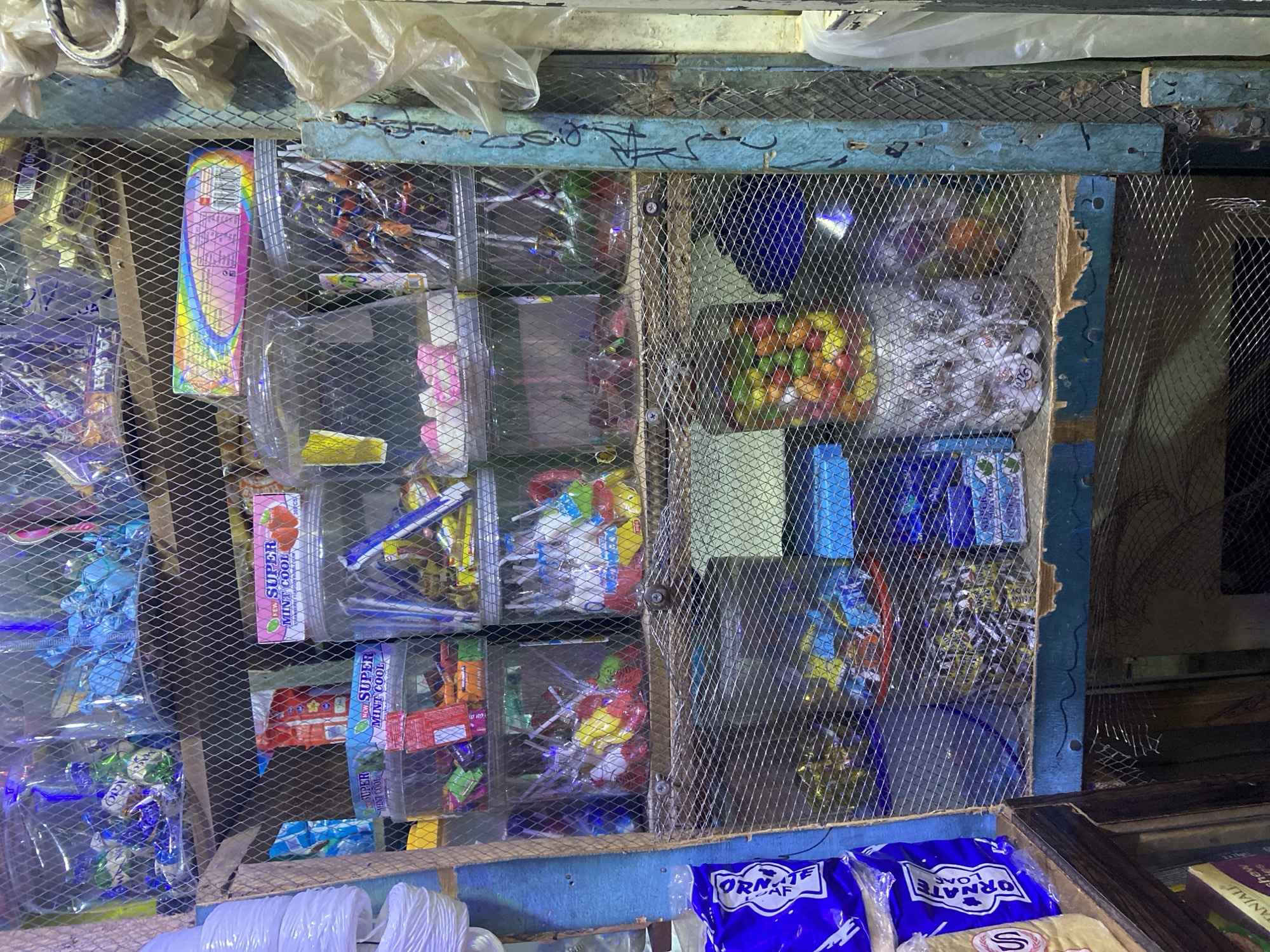 The image depicts a small, cluttered shop in Du, Nigeria. The shop is filled with various sweets and candies displayed in clear plastic containers and bags on shelves. The shelves are made of wood and metal mesh, and the entire display is enclosed by a wire mesh fence.
In the foreground, there are several blue plastic bags with white lettering that appears to say "ORNATE". Behind these, a variety of candies are visible, including lollipops in different colors and flavors, wrapped candies in colorful packaging, and small, individually wrapped sweets. Some of the candies are in large, clear plastic jars, while others are in smaller bags or boxes.
In the background, more shelves are filled with an assortment of sweets. Some of the packaging is in a language that appears to be Arabic, and some of the candies have Arabic writing on them. There are also some brightly colored packets of sweets, including one with a rainbow design.
The shop appears to be dimly lit, with some artificial lighting casting a glow on the candies. The overall impression is one of abundance and variety, with a focus on colorful and sweet treats.