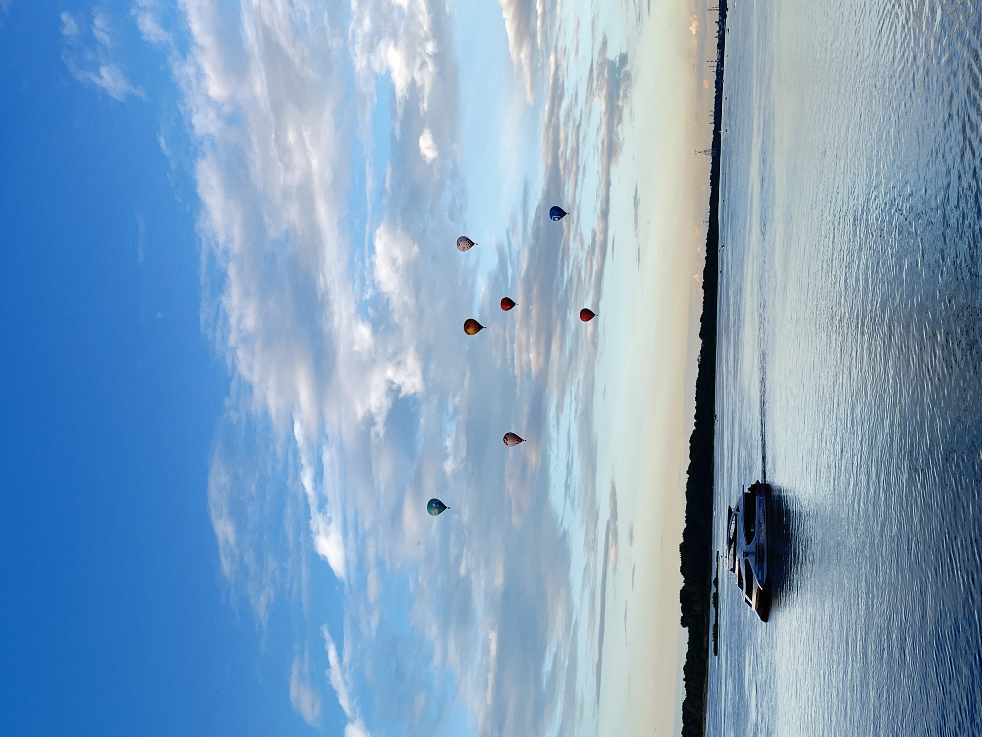 The image captures a scenic view of a wide river with a boat sailing on its surface. The river is vast and calm, reflecting the sky above. On the left side of the image, the sky is a vibrant blue, dotted with fluffy white clouds that stretch across the frame. The light suggests it is either early morning or late afternoon, with the sun casting a warm glow on the horizon. Several hot air balloons are visible in the sky, adding a touch of color and whimsy to the scene. They appear to be engaged in a ballooning activity, possibly a festival or recreational flight.
On the right side of the image, the river occupies most of the space, its surface rippling gently. A dark silhouette of trees lines the far bank of the river, creating a distinct separation between the water and the land. A boat is prominently featured on the river, its dark hull contrasting with the lighter tones of the water. The boat appears to be moving, leaving a subtle wake behind it. In the distance, along the horizon, some structures are faintly visible, hinting at a populated area or industrial presence. The overall mood is peaceful and serene, with a sense of open space and natural beauty. The image captures a harmonious blend of natural elements – sky, water, and land – with human-made objects like the boat and hot air balloons. There is no visible text in the image.