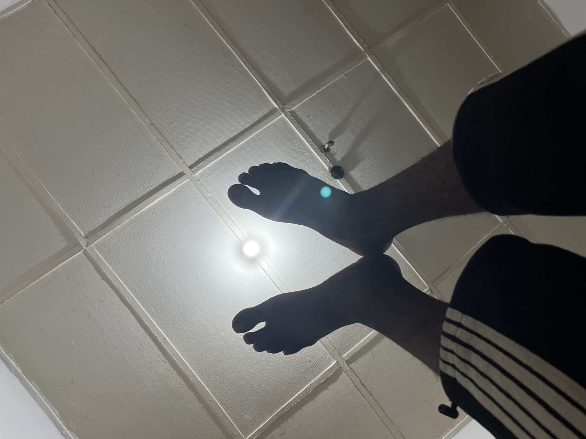 An extreme low-angle, wide shot shows two bare feet and lower legs from the perspective of someone lying on their back, looking up at the ceiling. The feet are silhouetted against a bright light source, possibly a ceiling light fixture, which creates lens flare. The ceiling is white and divided into square panels by thin, raised lines. The person is wearing dark pants with thin white stripes on the lower part of one leg, visible in the right side of the frame. The feet are positioned in the lower half of the frame, with the upper feet and legs extending out of view.