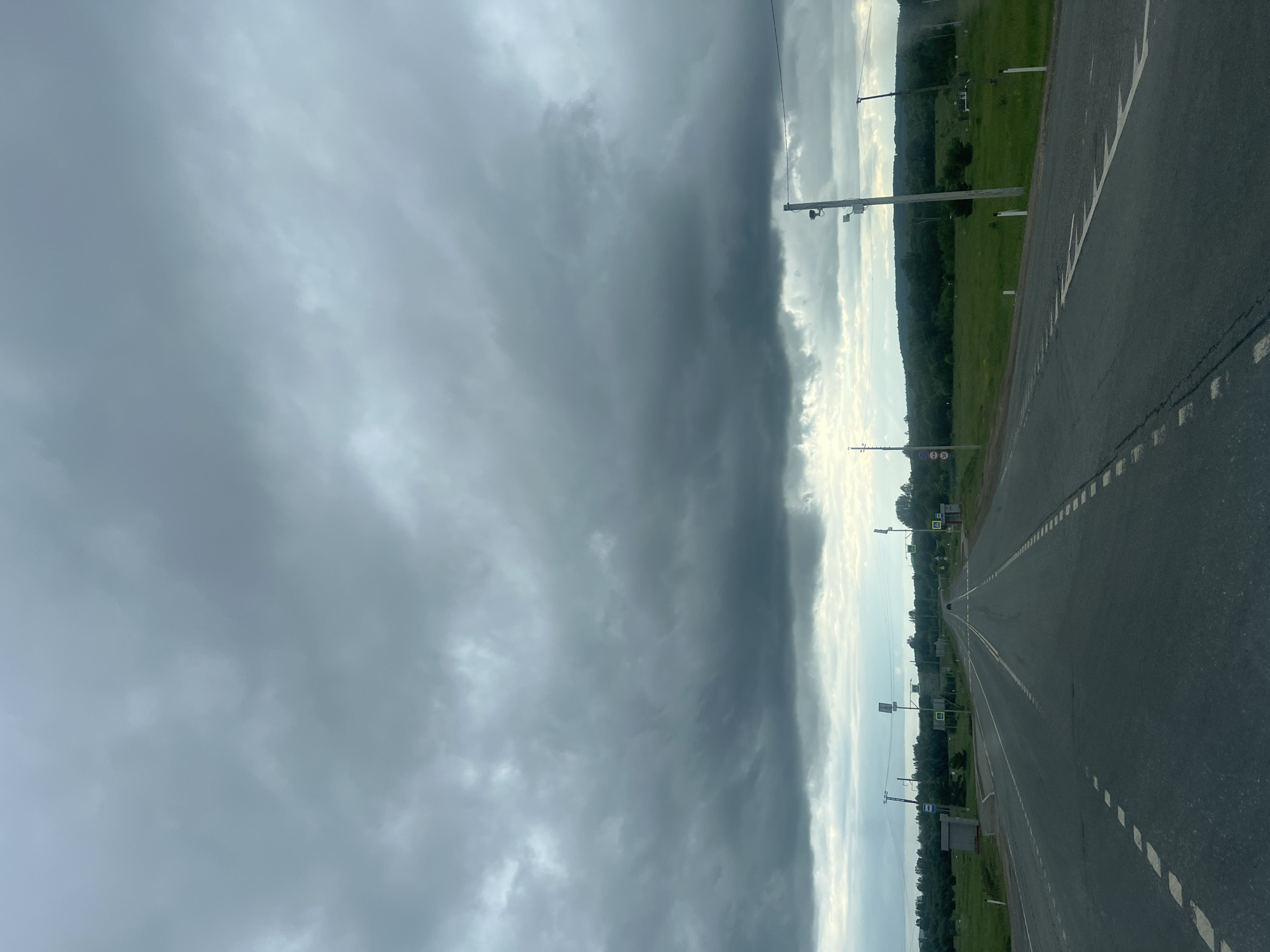 The image captures a view from a vehicle traveling on a paved road in Гремяча, Russia. The road is asphalt with white dashed lines indicating lanes and is bordered by grassy shoulders. The surrounding landscape is rural, with green fields and a dense line of trees in the distance, forming a horizon. The sky is heavily overcast with dark, heavy clouds, suggesting impending rain or stormy weather. Several utility poles with electrical wires and street lights are visible along the roadside. Road signs are present, including what appear to be pedestrian crossing signs and speed limit signs. A small bus stop shelter is also visible on the left side of the road. No people or animals are clearly discernible in the scene, and the overall atmosphere is somber due to the weather conditions.
