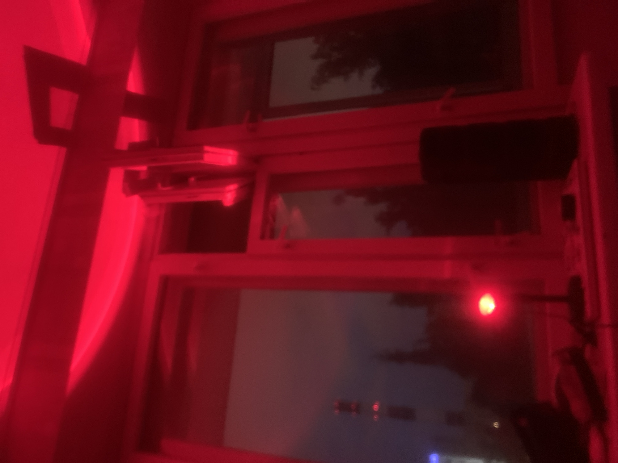 The image displays an indoor scene bathed entirely in red light, looking out through a multi-pane window during what appears to be night or late dusk. The window frame, likely made of a light-colored material, is saturated with the intense red illumination.
On the left side of the window, a complex, L-shaped mounting bracket, possibly metallic, supports several stacked electronic devices, which could be sensors, small computers, or other equipment. These devices also appear reddish due to the lighting. To the right, mounted horizontally on the window frame, is a dark, rectangular object, possibly a speaker or another electronic component. Further to the right and lower, a single, bright red LED or indicator light glows, attached to a slender arm.
Outside the window, the view is dark and somewhat blurry, showing indistinct shapes resembling trees or foliage against a dimly lit sky. Faint lights are discernible in the distance through the lower window pane, possibly indicating a distant city or residential area, or reflections. On the far left, folds of a red curtain or drape are visible, blending with the overwhelming red hue of the scene. The pervasive red light creates a distinct, atmospheric, and possibly functional setting, such as a darkroom, security area, or a space with specialized lighting. No text is visible in the image.