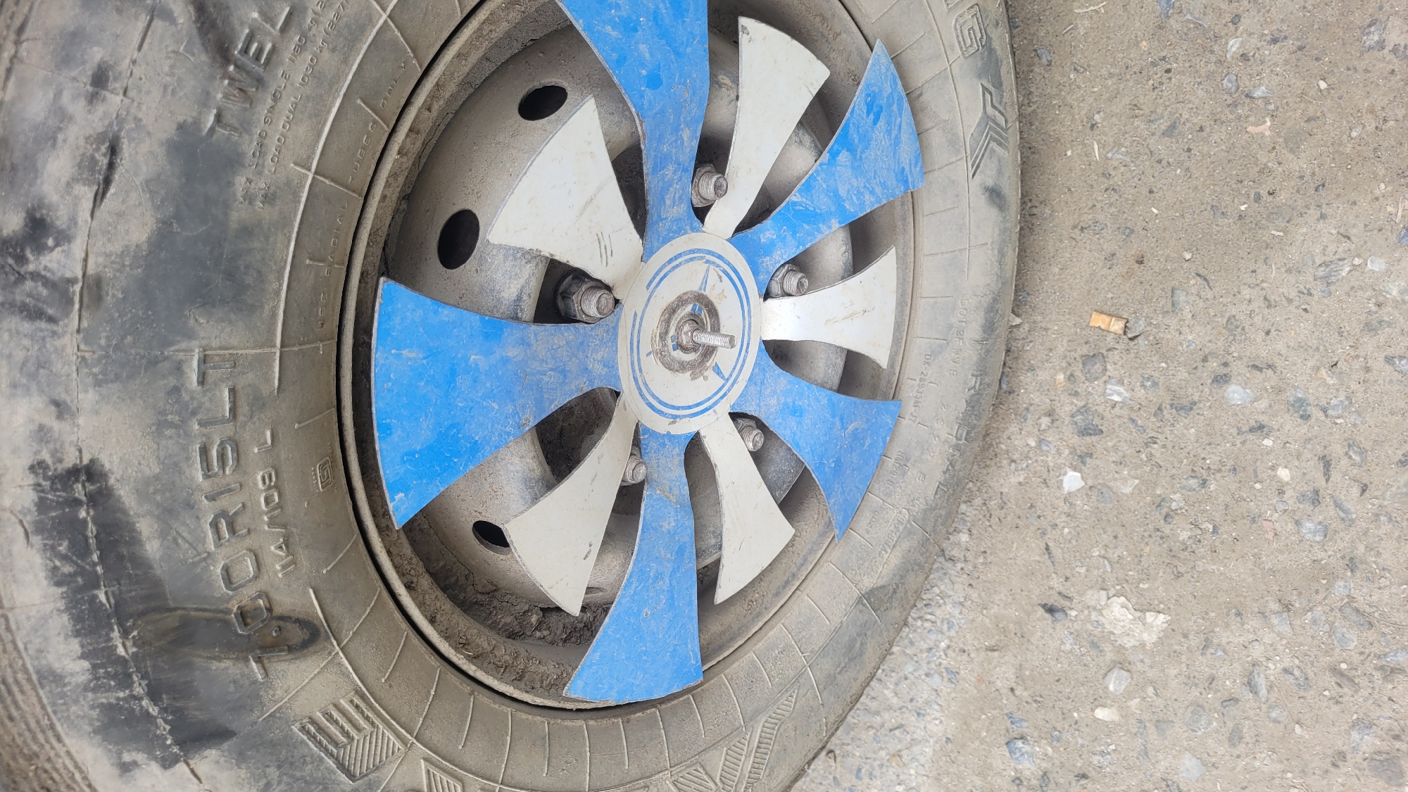 A close-up, eye-level shot shows the front of a blue and white fan positioned over the wheel of a vehicle. The fan's blades are a mix of white and blue, attached to a central silver hub. The vehicle's tire, visible around the fan, is black and appears worn, with dust and dirt on its surface. Markings on the tire are partially obscured by the fan and dirt, but "7.00R15LT" and "TWEL" are legible. The background is a rough, gray, and gravelly surface, possibly a road or ground in Vashisht, India, with a discarded cigarette butt visible in the lower right quadrant of the image. The overall lighting is natural, suggesting an outdoor environment.