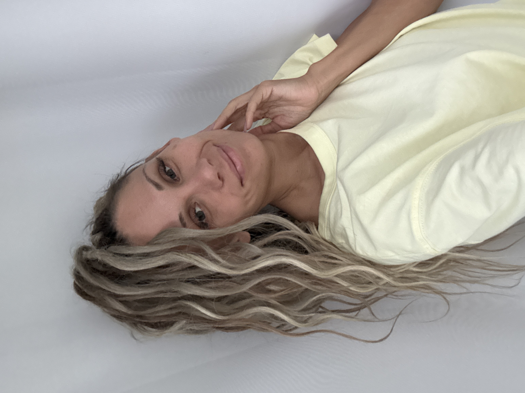 A woman with blonde dreadlocks pulled into a high ponytail smiles as she rests her hand on her cheek. She is wearing a light yellow t-shirt and is against a white wall with vertical folds. The image appears to be a selfie taken in the city of Rusanovo, Russia.