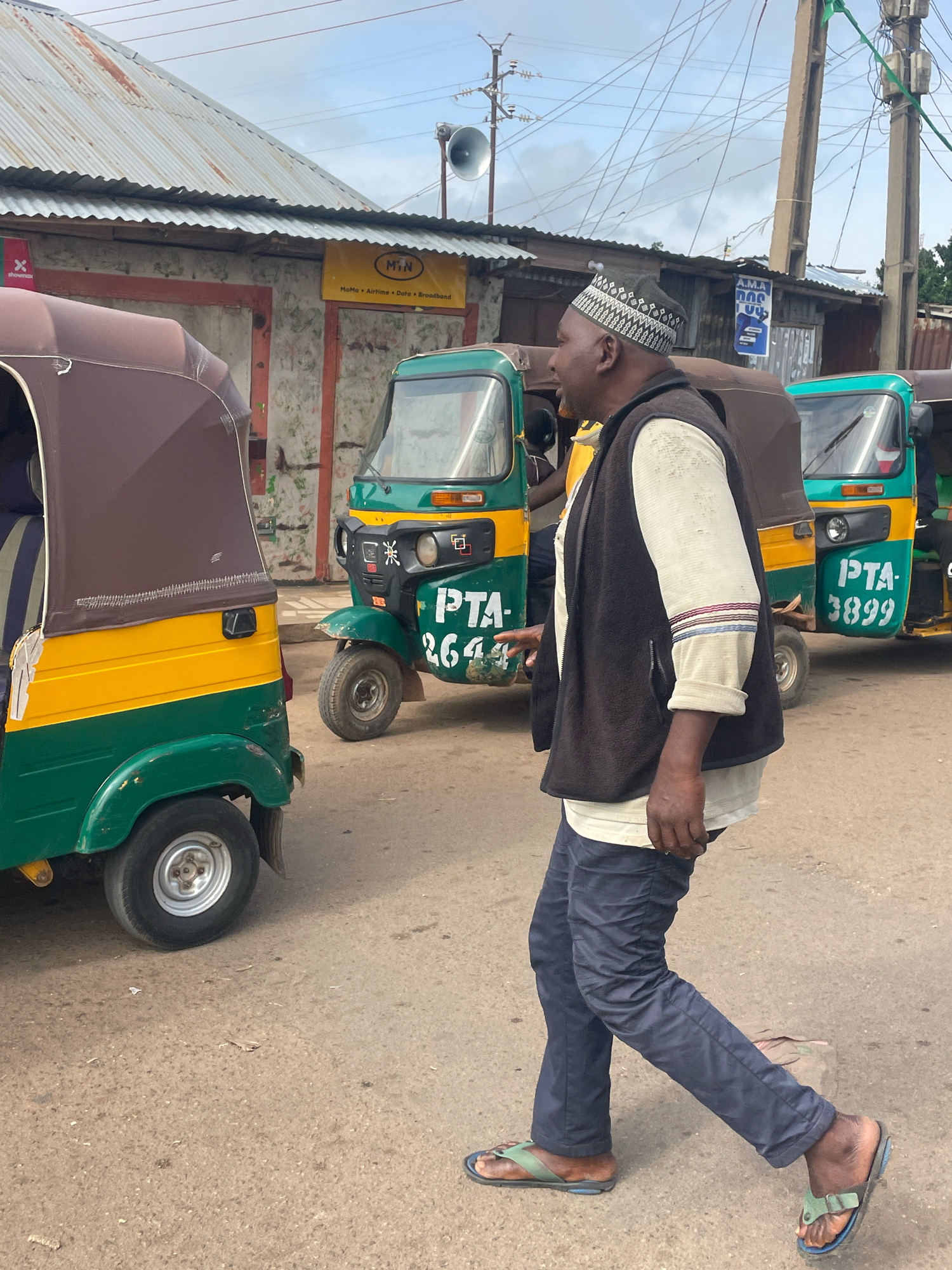 A man wearing a black patterned kufi, a light-colored long-sleeved shirt, a dark vest, and dark blue pants walks on a paved road in front of several auto-rickshaws. The auto-rickshaws are green and yellow. One auto-rickshaw in the foreground on the left has the license plate "PTA 2646". Another auto-rickshaw to the right has the license plate "PTA 3899". Behind the auto-rickshaws, there is a building with a corrugated metal roof and a weathered facade. A yellow sign with black text that reads "MIN" and "MoMa Airtime. Data. Broadband" is visible on the building. Electrical wires and utility poles are visible in the background. The sky is cloudy. The scene appears to be in a market or busy street in Gura Top, Nigeria.