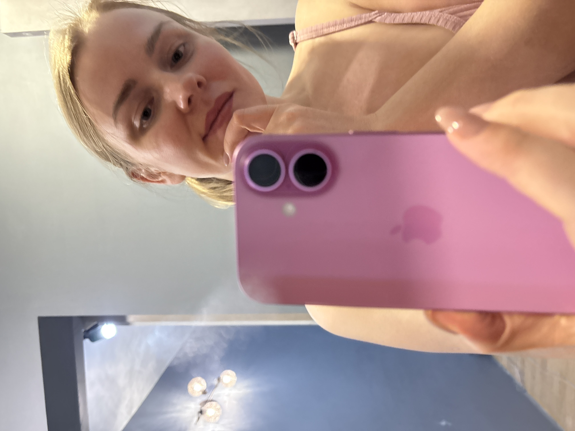 A young, fair-skinned woman with blonde hair is taking a selfie indoors with a pink smartphone. She is wearing a light pink top. The background shows a wall with a light fixture. It's not possible to verify the location as Chaikovsky, Russia based on the visual information.