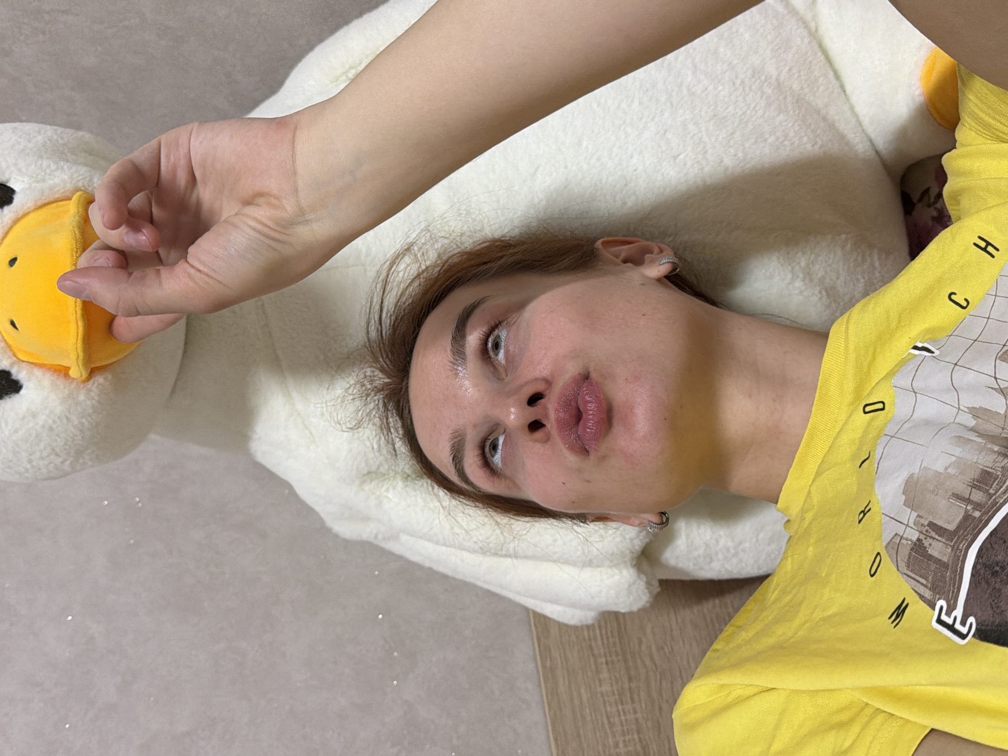 A woman lies on her back on a white, fluffy surface, possibly a large stuffed animal, wearing a yellow t-shirt with a graphic print. Her head rests against the white material, and she is looking up towards the camera with a neutral expression. Her complexion is fair, and her hair is light brown, parted slightly to one side. An arm and hand reach into the frame near the plush toy, which features a yellow beak. The background includes a light grey surface and a wooden-like surface. The city of Chaikovsky, Russia, cannot be determined from the visual information provided.
