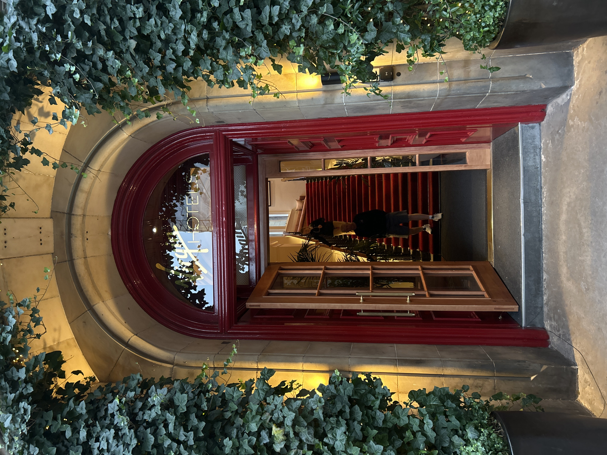 The image captures the entrance to The Raeburn, a hotel located at 132 Dundas Street, City of Edinburgh, United Kingdom. The entrance features a grand, arched doorway framed by ivy-covered stone pillars. The doorway itself is adorned with a deep red frame, with a curved glass panel above displaying the hotel's name in a stylized gold font. To the left and right of the entrance, large potted plants with dense green foliage add to the sophisticated atmosphere.
The double doors of the entrance are open, revealing a red carpeted staircase leading upwards. A person, wearing a dark jacket and light-colored shorts, is walking up the stairs, viewed from the back. Another person, wearing a dark top and light-colored shorts, is standing further up the stairs, also facing away from the camera. Inside the entrance, glimpses of palm plants and a cozy seating area can be seen. The lighting suggests it is evening or dusk, with warm yellow lights illuminating the stone archway and exterior walls. The ground in front of the entrance is paved, with a dark mat laid at the threshold of the doorway.
