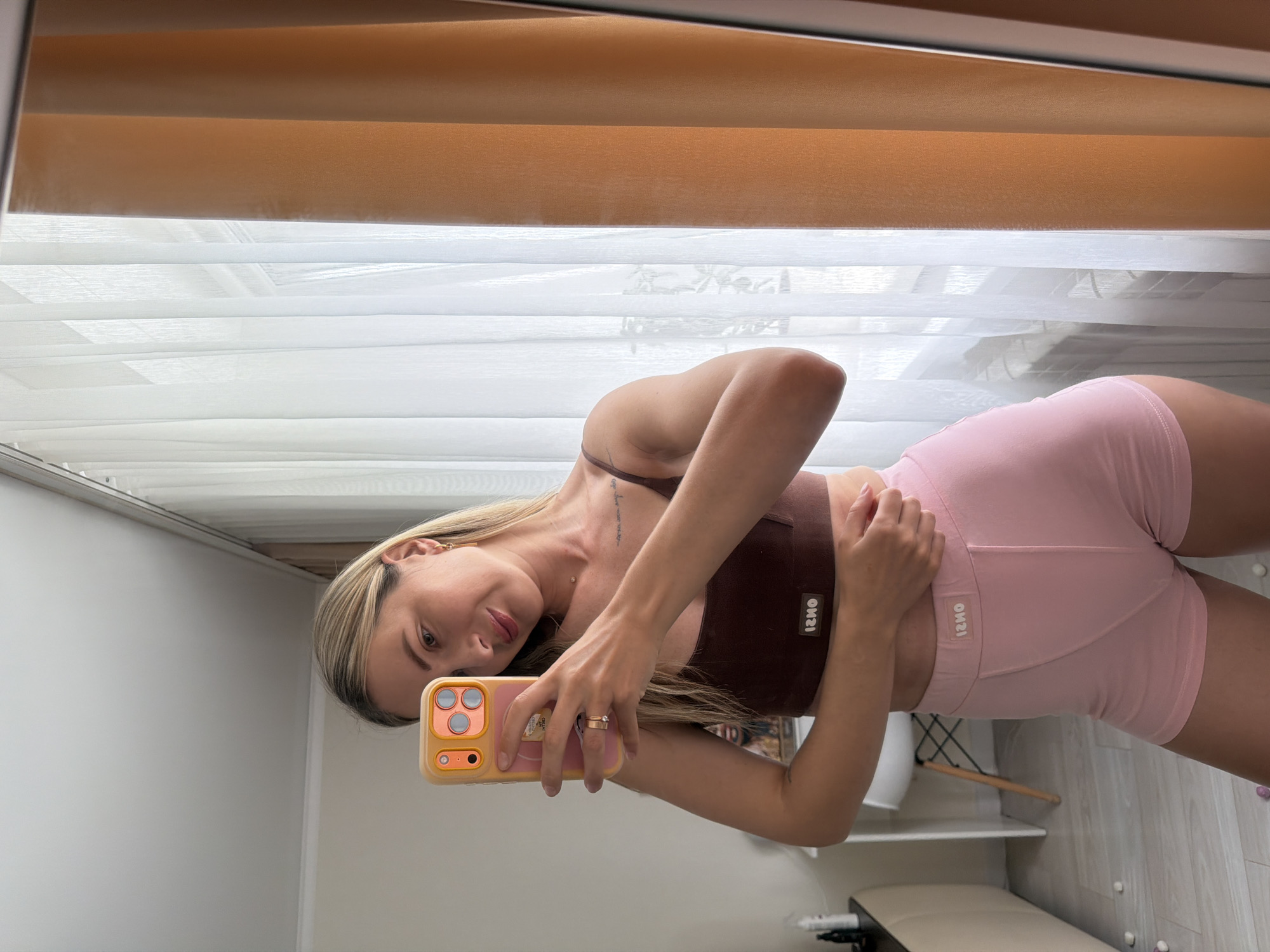 A blonde woman with fair skin and light makeup takes a selfie in a mirror. She is wearing a brown bralette and pink shorts. She has a ring on her left hand and a bar-shaped earring in her right ear. Her hair is long and blonde, pulled back from her face. She is smiling slightly.
The woman is standing in what appears to be a bedroom or dressing area. Behind her, a sheer white curtain is visible, with an orange vertical blind partially obscuring the view. A radiator is visible beneath the window. To her left, a white chair and a small table with bottles on it can be seen. The floor is light wood. The overall lighting suggests it is daytime.
This image does not contain any identifiable landmarks or features that would allow for a specific location within Semiluki, Russia, to be determined.