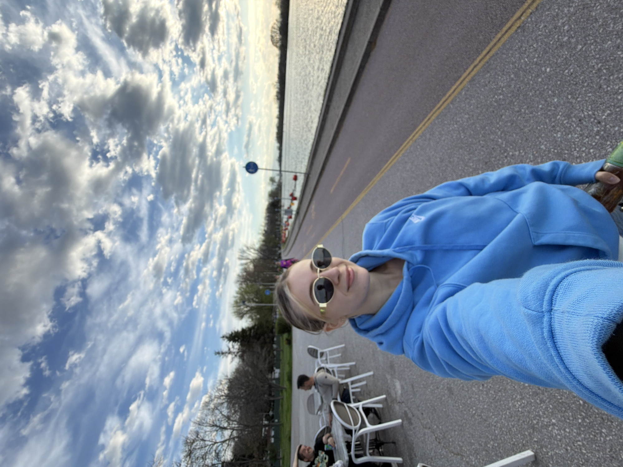A young woman with blonde hair pulled back in a bun is taking a selfie while wearing round sunglasses and a light blue hooded sweatshirt. She is standing on a grey paved area next to a body of water. The weather appears to be sunny with a partly cloudy sky. To the left are trees and a row of white chairs and tables where at least two people are seated. Behind the paved area and chairs is a grassy area. The background includes a road that runs along the edge of the water. It is not possible to determine the location in Plovdiv, Bulgaria, from the image.