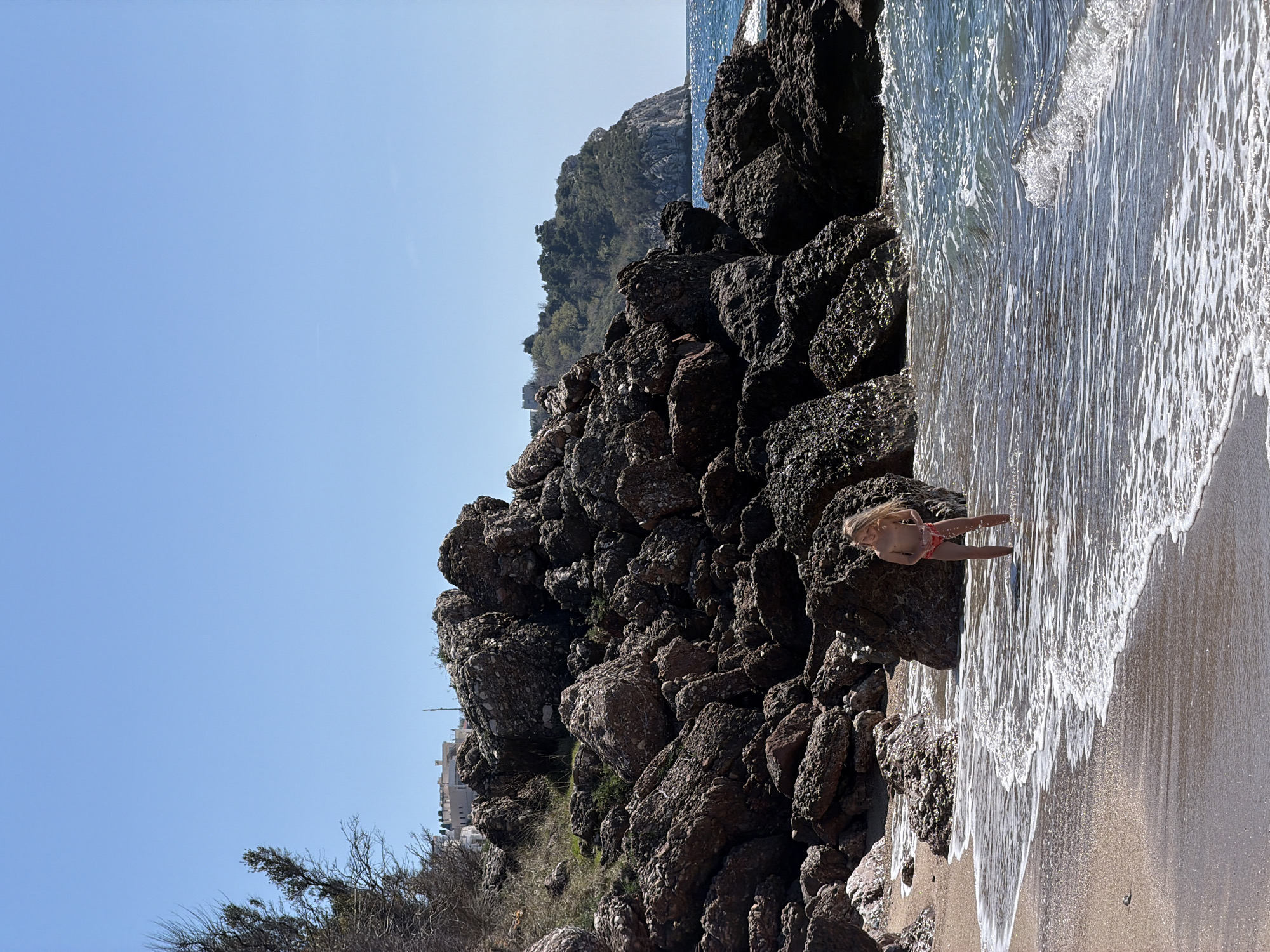 A scenic, sunlit beach scene in Drobnići, Montenegro, presents a rocky shoreline juxtaposed against the gentle wash of the sea. A young girl with blonde hair stands near the edge of the water, her figure small against the broader landscape. The beach consists of a sandy area, partially covered by the retreating waves, alongside a jumble of dark, large rocks. The background includes a hillside densely covered with vegetation. The sky is clear and blue, completing the coastal setting.