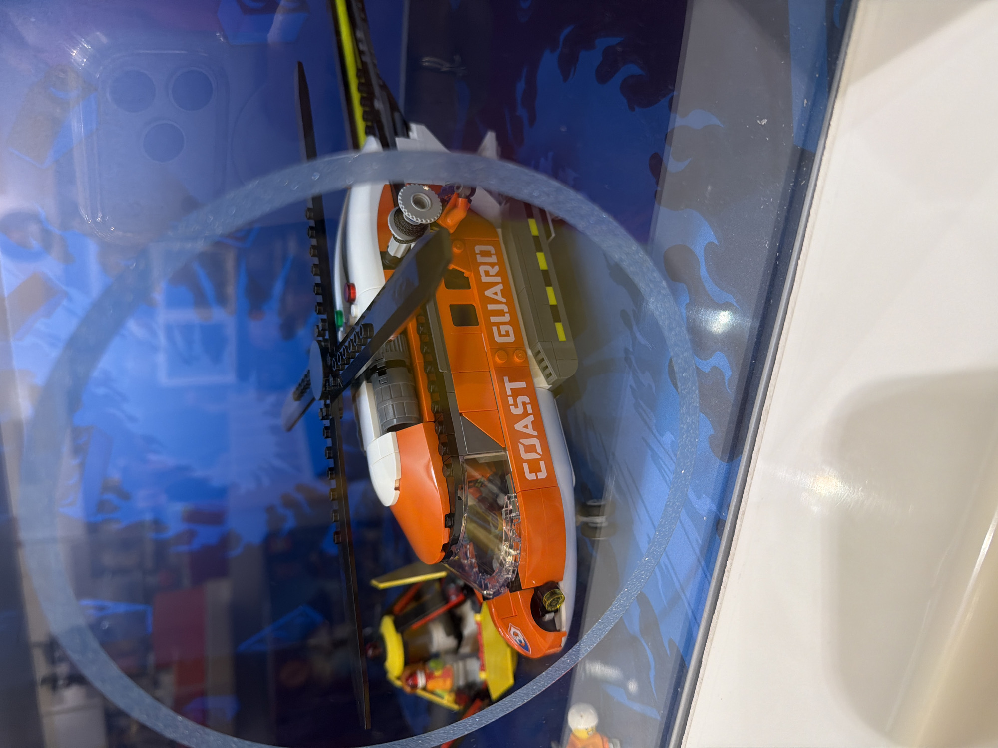 A LEGO Coast Guard helicopter is displayed inside a clear plastic box. The helicopter is predominantly orange and white, with "COAST GUARD" printed in white letters on the side. It has a black rotor and tail fin, and a clear cockpit. The background of the display box features a blue, wave-like graphic, suggesting a marine or water-related theme. The helicopter appears to be a model, possibly part of a toy set. The lighting suggests an indoor setting, likely a store or display case. There is a faint reflection on the plastic, showing what appears to be a phone case with three circular camera lenses. The image is oriented sideways.