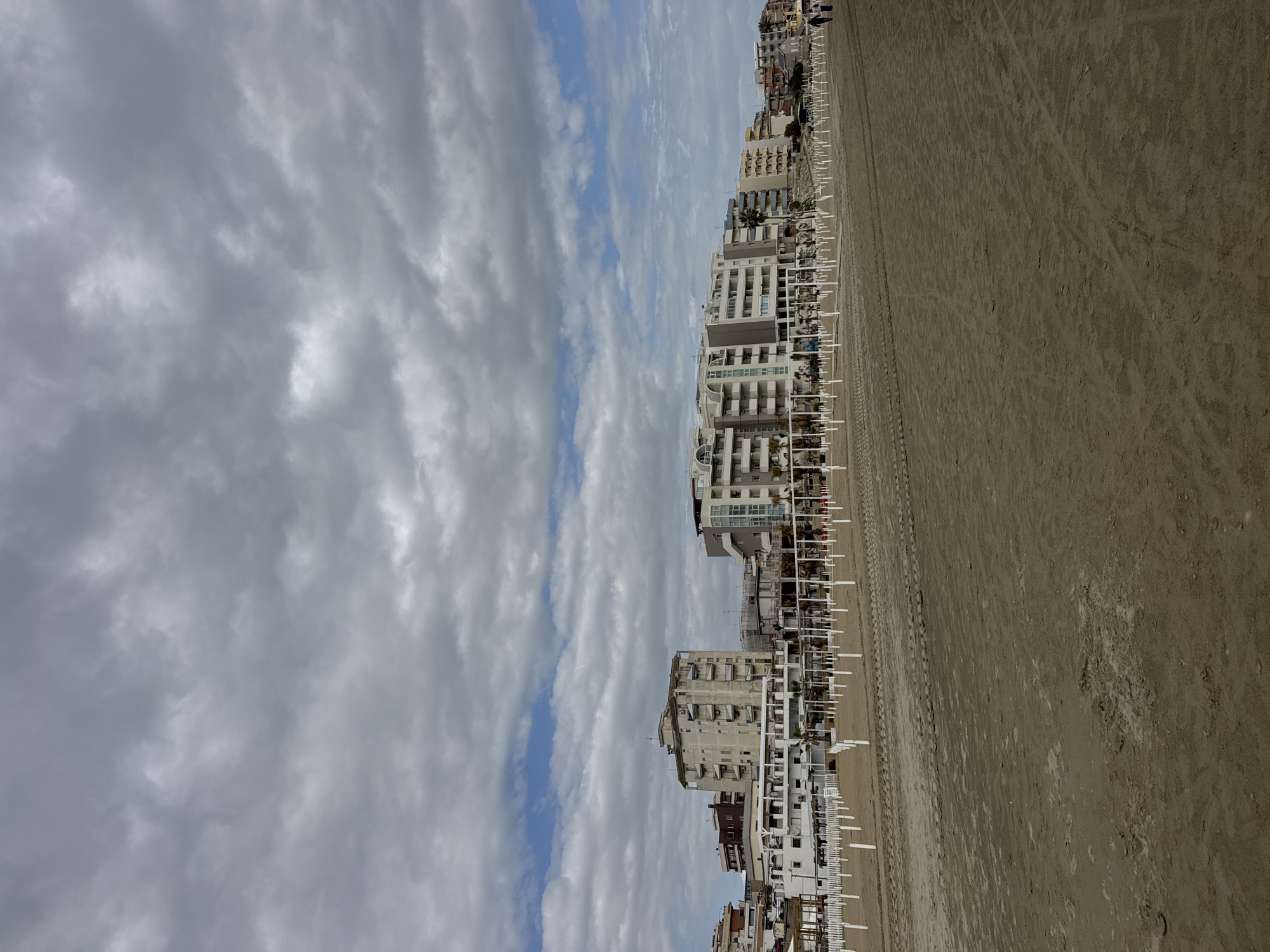 This image presents a wide view of a sandy beach lined with buildings, likely in a coastal town such as Nettuno, Italy. The scene appears calm and devoid of significant activity.
The upper portion of the frame is filled with a dynamic sky, characterized by a mix of grey and white clouds interspersed with distinct streaks of blue sky. This suggests a day with variable weather, possibly partly cloudy or transitioning between clear and overcast conditions.
Along the left side of the image, a continuous row of multi-story buildings fronts the beach. These structures, possibly hotels or apartment complexes, exhibit varied architectural styles but are generally light-colored with prominent balconies and windows. In front of these buildings and extending onto the sand are numerous evenly spaced white frameworks, which appear to be the supports or bases for beach umbrellas or sunbeds, typical of commercial beach establishments.
The right side of the image is dominated by a wide, flat expanse of light brown sand. The sand shows various textural patterns, including tracks and subtle undulations, but is otherwise empty.
No people or animals are clearly visible, and no specific activities are taking place on the beach or in the water (which is not visible). The overall impression is one of a beach either in the off-season, early in the morning, or late in the day, before or after peak activity. The lighting is diffused, consistent with the partly cloudy sky. There is no visible text in the image.