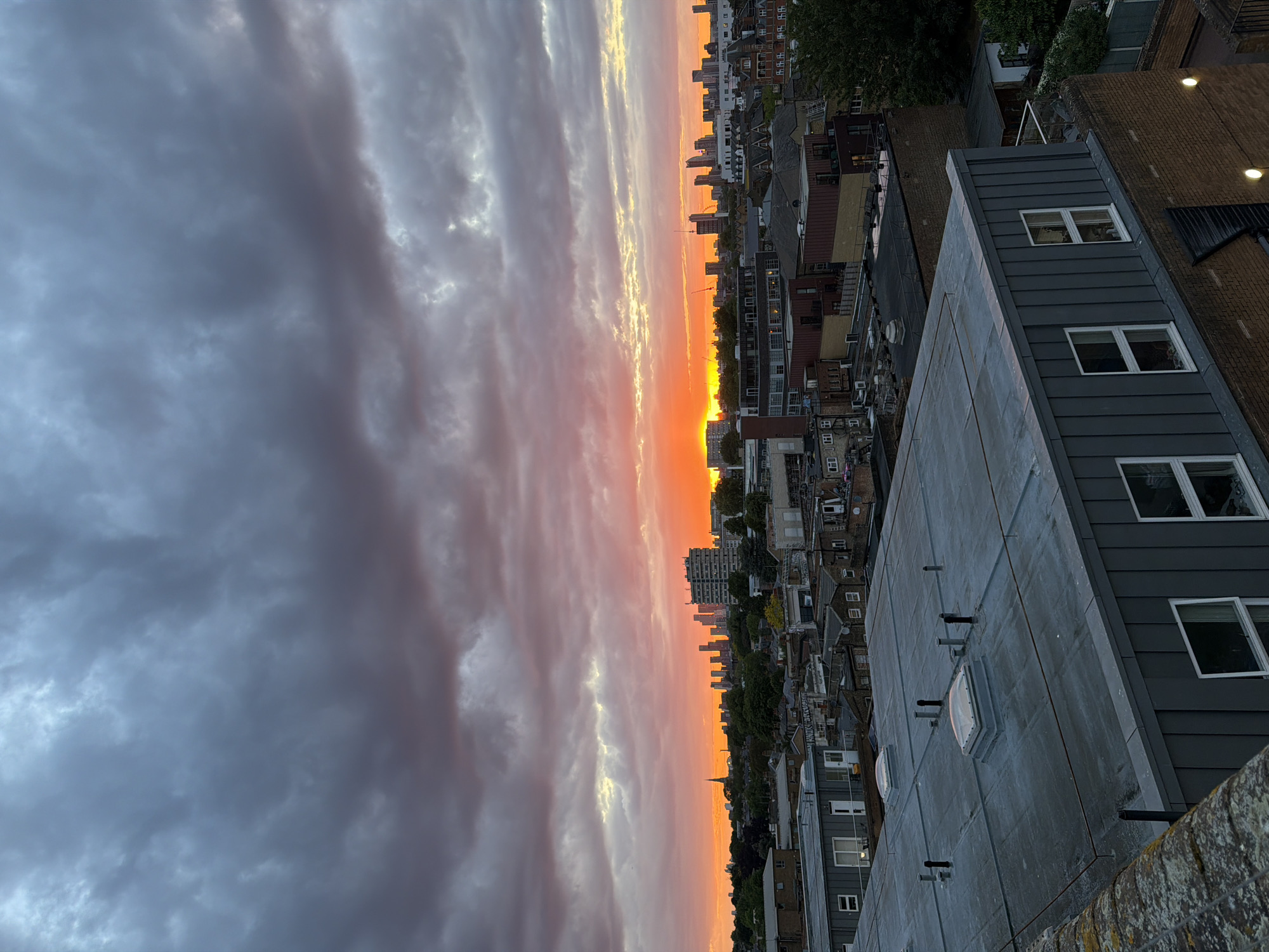 The image is a high-angle, slightly tilted view of a sunset over London, United Kingdom. The foreground features a flat, grey rooftop of a building with several skylights and ventilation pipes. In the midground, a dense collection of residential and commercial buildings stretches towards the horizon, interspersed with green trees. The background showcases a vibrant sunset, with a gradation of colors from dark grey clouds in the upper left to a bright orange and yellow sky at the horizon line. The skyline is punctuated by a few taller buildings and the silhouette of what appears to be the London Eye.