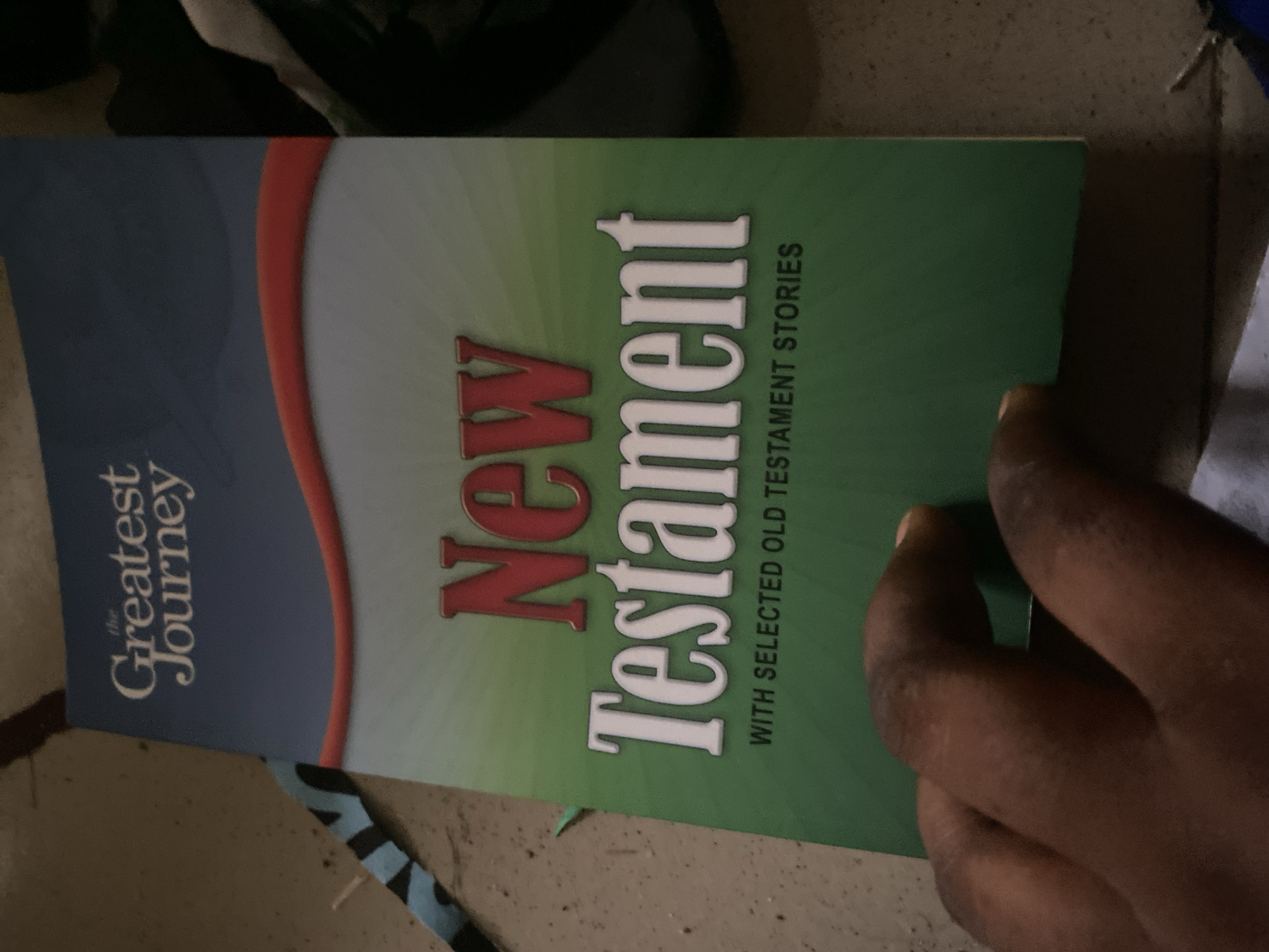 A book titled "The Greatest Journey: New Testament with Selected Old Testament Stories" is displayed at an angle. The book has a dark blue upper section with white text and a red curved line separating it from a green section below. The words "New" are in red, and "Testament" is in large white letters. Beneath this, in smaller black text, is "WITH SELECTED OLD TESTAMENT STORIES". A person's dark-skinned hand is visible in the lower left corner, obscuring part of the book. The book rests on a surface that appears to be a floor or ground, with scattered dirt and debris. A black and blue striped object is partially visible to the left of the book. The lighting is dim, suggesting an indoor or shadowed environment. The scene is located in Jauro Yinu, Nigeria.