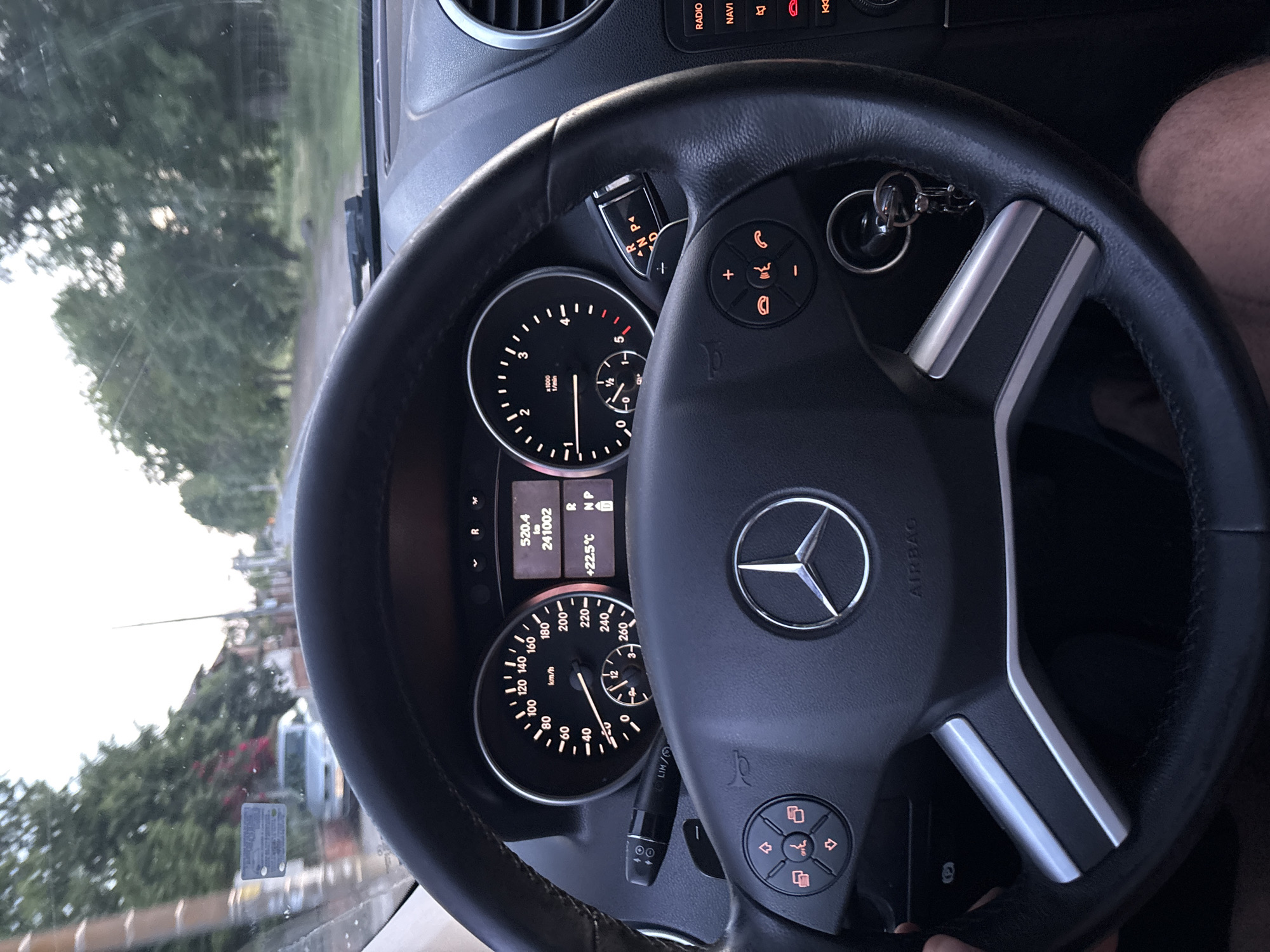 The image shows the interior of a Mercedes-Benz car, focusing on the dashboard and steering wheel. The odometer reading is 241002 km, and the outside temperature is +22.5°C. The speedometer shows a reading of 0 km/h, suggesting the vehicle is stationary. The car key is in the ignition. The steering wheel has several buttons for controlling various car functions, and the Mercedes-Benz logo is prominently displayed in the center. Outside the windshield, a blurred view of trees and buildings indicates the car is likely on a city street.
