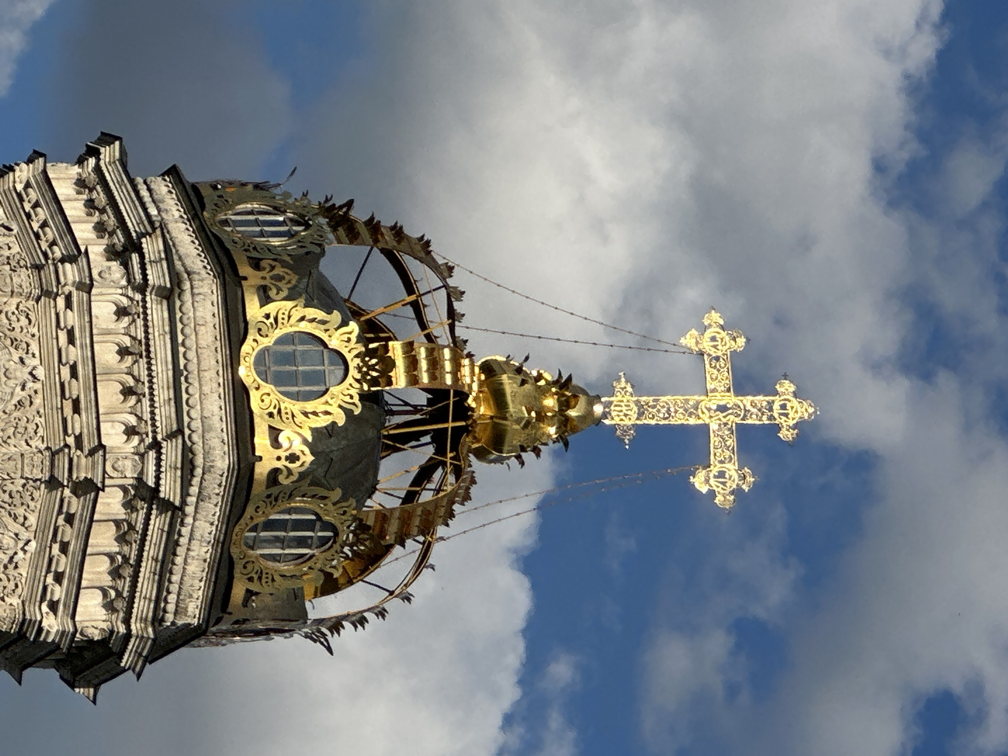The image shows a close-up of the gilded dome and cross of the Church of the Nativity of the Blessed Virgin Mary, located in the village of Dubrovitsy, Russia. The church's architecture is Baroque, characterized by intricate stone carvings and ornate gilded elements. The dome is a dark color, possibly dark green or black, topped with a circular open structure adorned with decorative gold elements. Above this structure, a large, ornate gilded cross stands prominently against a blue sky with scattered white clouds. The cross is highly detailed, with decorative patterns and smaller cross elements at its extremities. The lower portion of the image reveals the carved stonework of the church's tower, featuring multiple layers of decorative moldings and sculpted figures or reliefs. The lighting suggests it is daytime, with sunlight illuminating the gilded elements and casting some shadows.