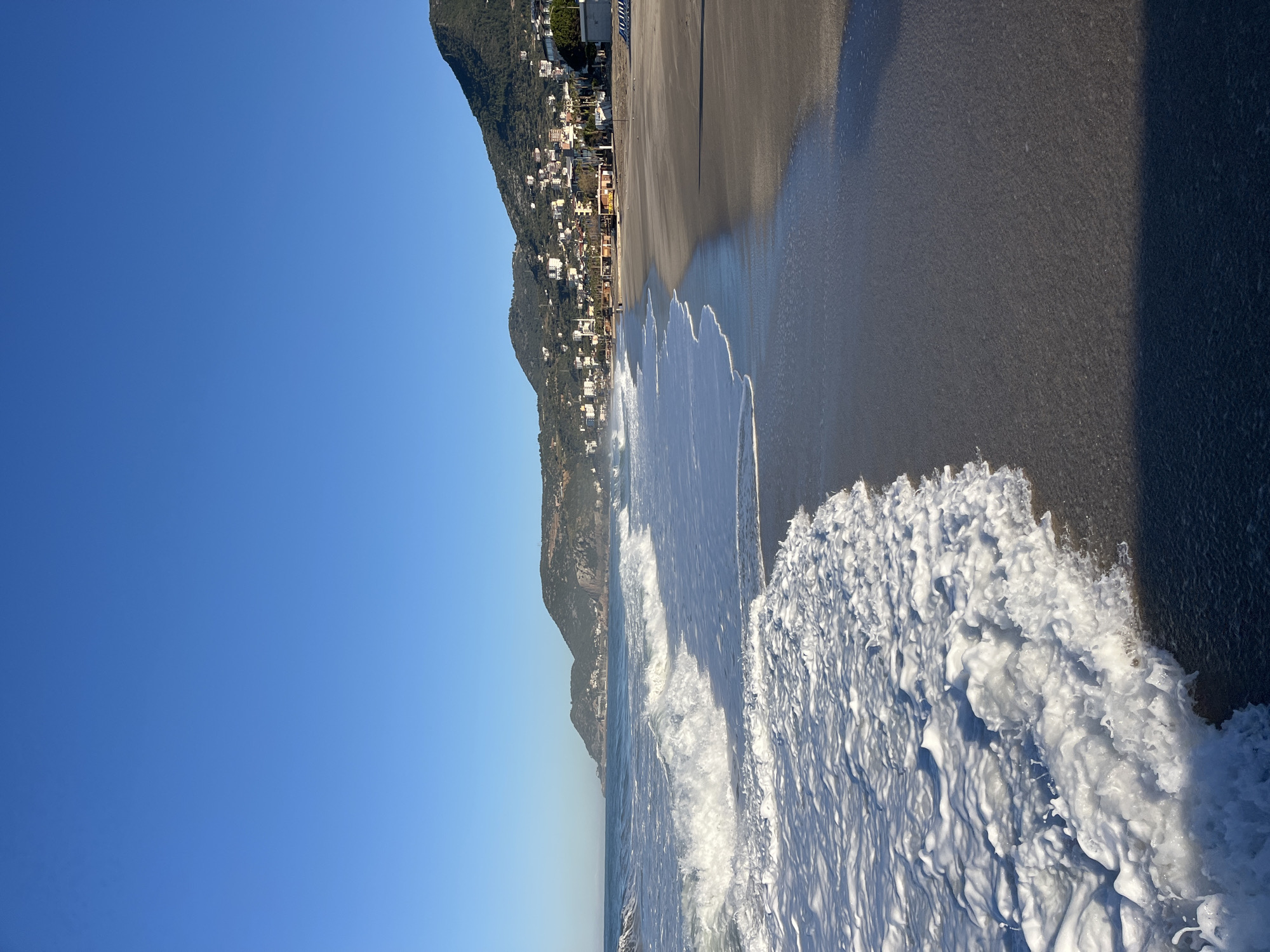 The image depicts a beach scene in Alanya, Turkey. The turquoise sea, churning with white foam, meets a dark sand beach. The coastline is lined with buildings and vegetation that are situated on a hill in the background. The sky is clear and blue.
