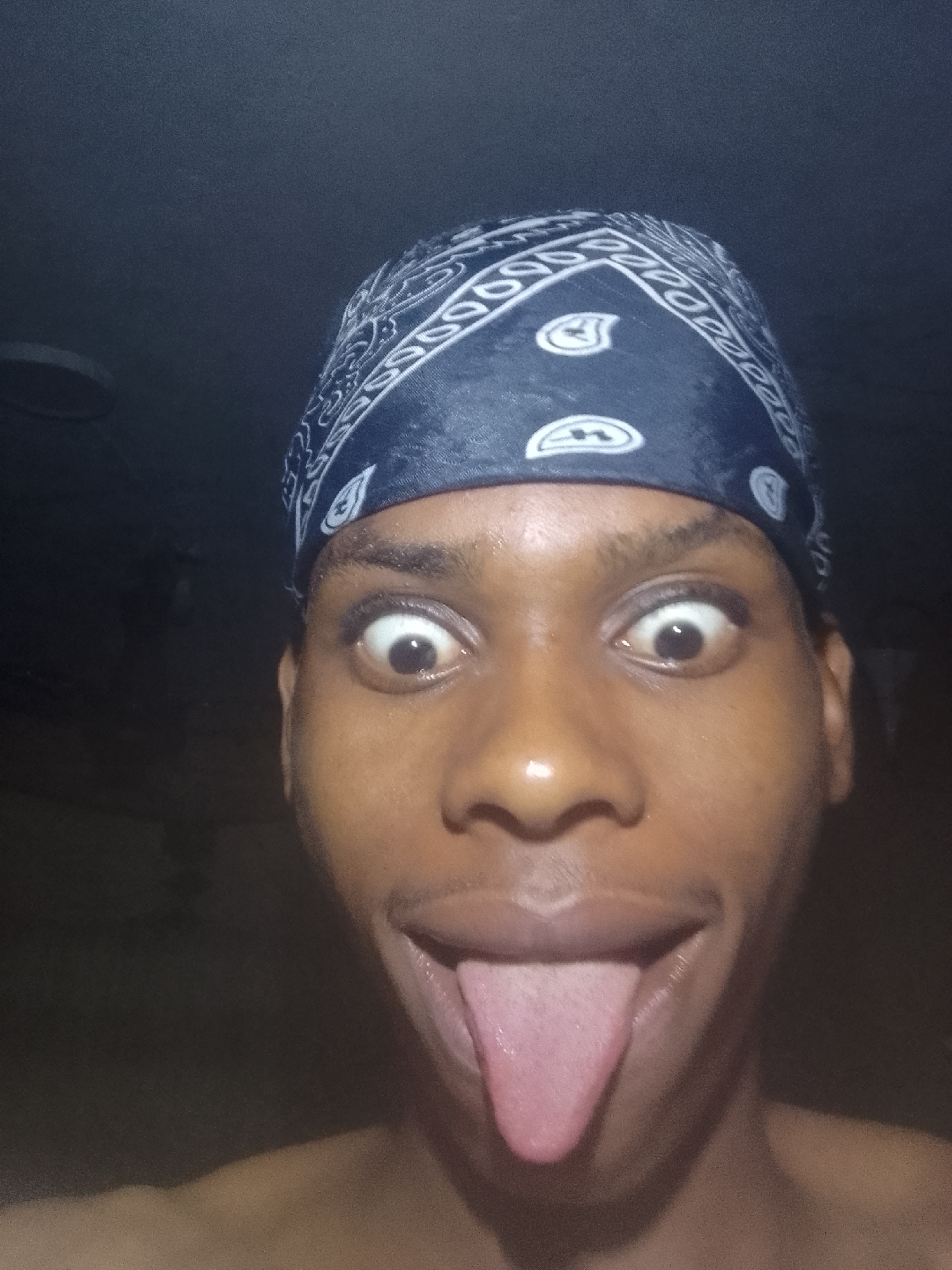 The image is a close-up, selfie-style photograph of a person with dark skin. The person is wearing a dark blue bandana on their head with a white paisley pattern. Their eyes are wide open, and they are sticking their tongue out. The background is dark and out of focus. The location is stated to be Nkpor, Nigeria, but there is no visual confirmation of the location in the image.