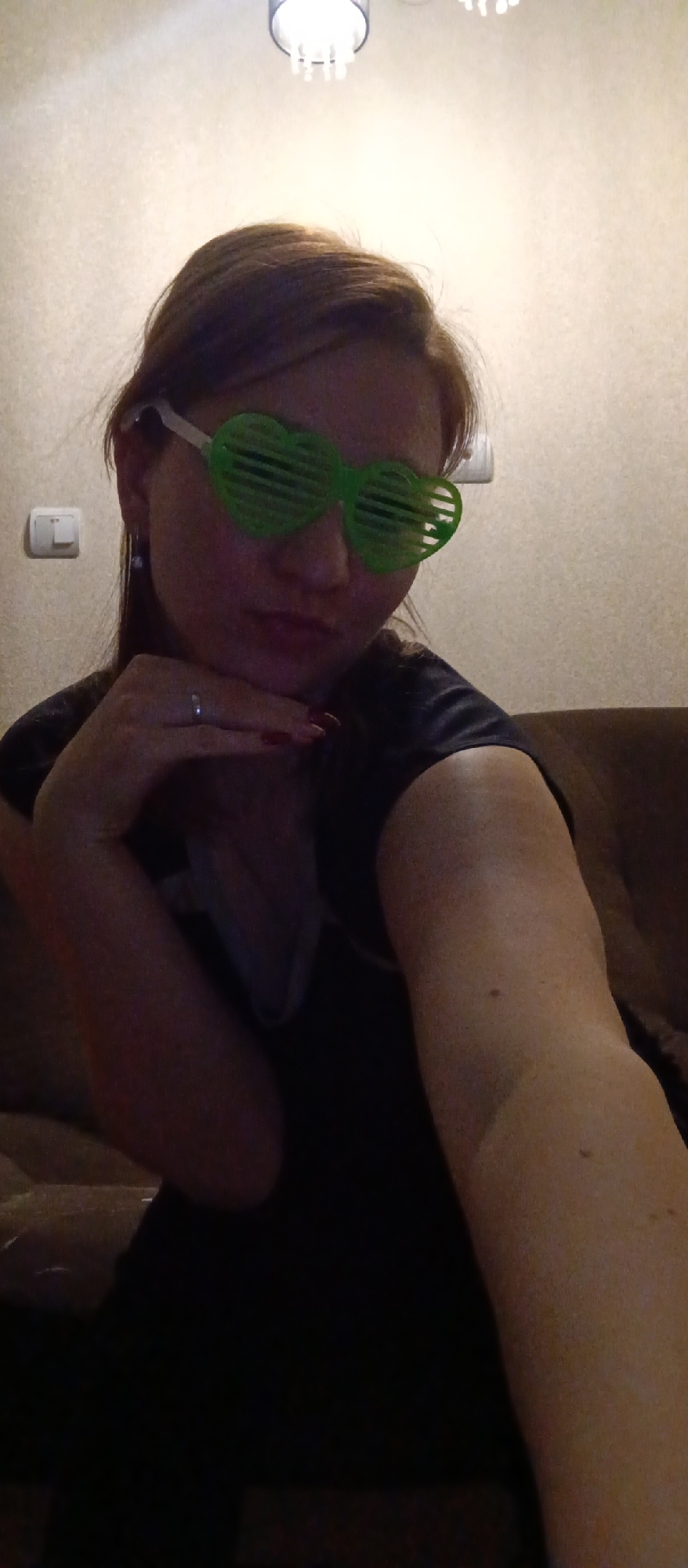 A woman is taking a selfie indoors, seated on a brown couch in an unknown location in the city of Unknown, Unknown. She is wearing green heart-shaped glasses and a dark top. Her left hand, adorned with a ring and painted fingernails, rests against her face. Behind her is a beige-colored wall with a light switch and a ceiling light.