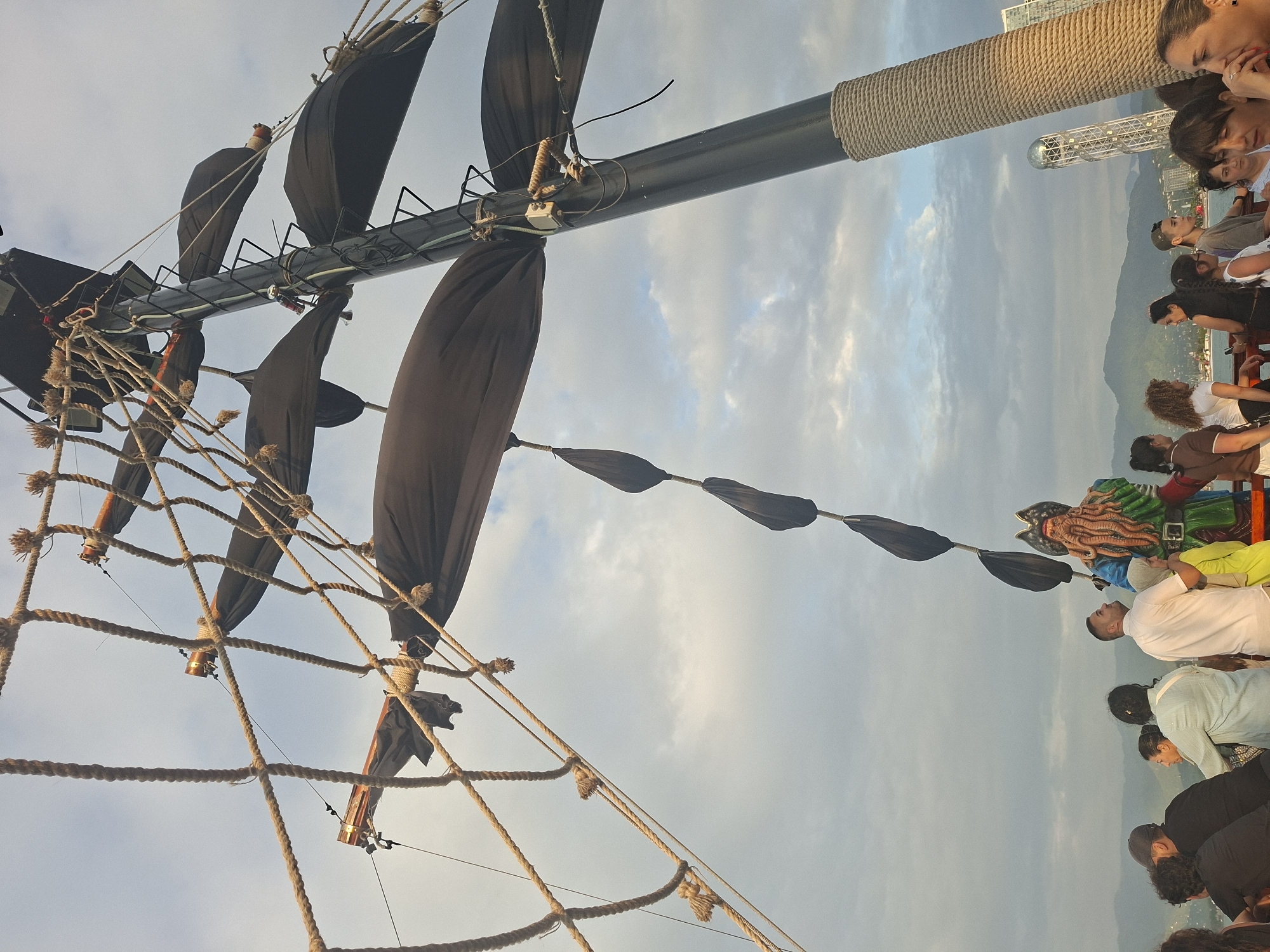 The image captures a view from a pirate-themed ship, likely on a tour, with a large mast and rigging dominating the foreground against a cloudy sky. Several dark, sail-like structures are attached to the mast. A costumed figure resembling a pirate with a long, orange beard and a green coat stands prominently on the deck, facing away from the camera. A group of diverse individuals, dressed in casual clothing, are gathered on the deck, some looking at the pirate figure, others at the scenery. In the background, a cityscape with mountains and a distinct tower structure, possibly the Alphabet Tower, is visible, suggesting the location is Batumi, Georgia. The overall scene suggests a recreational activity, possibly a tourist attraction, set against the backdrop of the city.