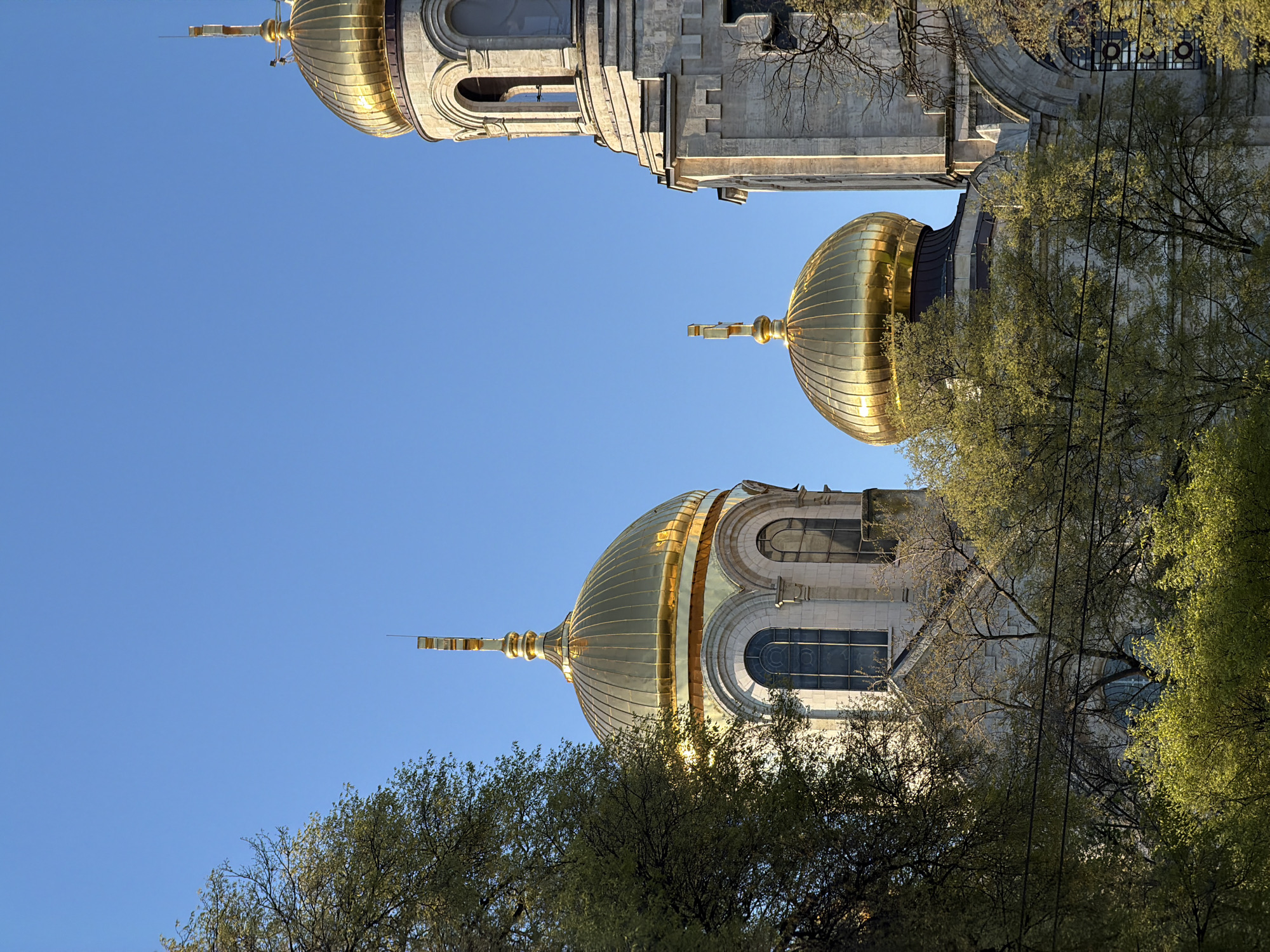 The image features the golden domes of the Dormition of the Mother of God Cathedral in Varna, Bulgaria, as seen from a low-angle perspective looking upward. The domes are arranged vertically in the frame, with the topmost dome cropped at the top. The domes are metallic and highly reflective, with visible seams in their construction. The building itself is visible below each dome, constructed of light-colored stone. Deciduous trees with green leaves partially obscure the lower domes and parts of the building. The sky is clear and blue, serving as a backdrop for the architecture. No people are visible.