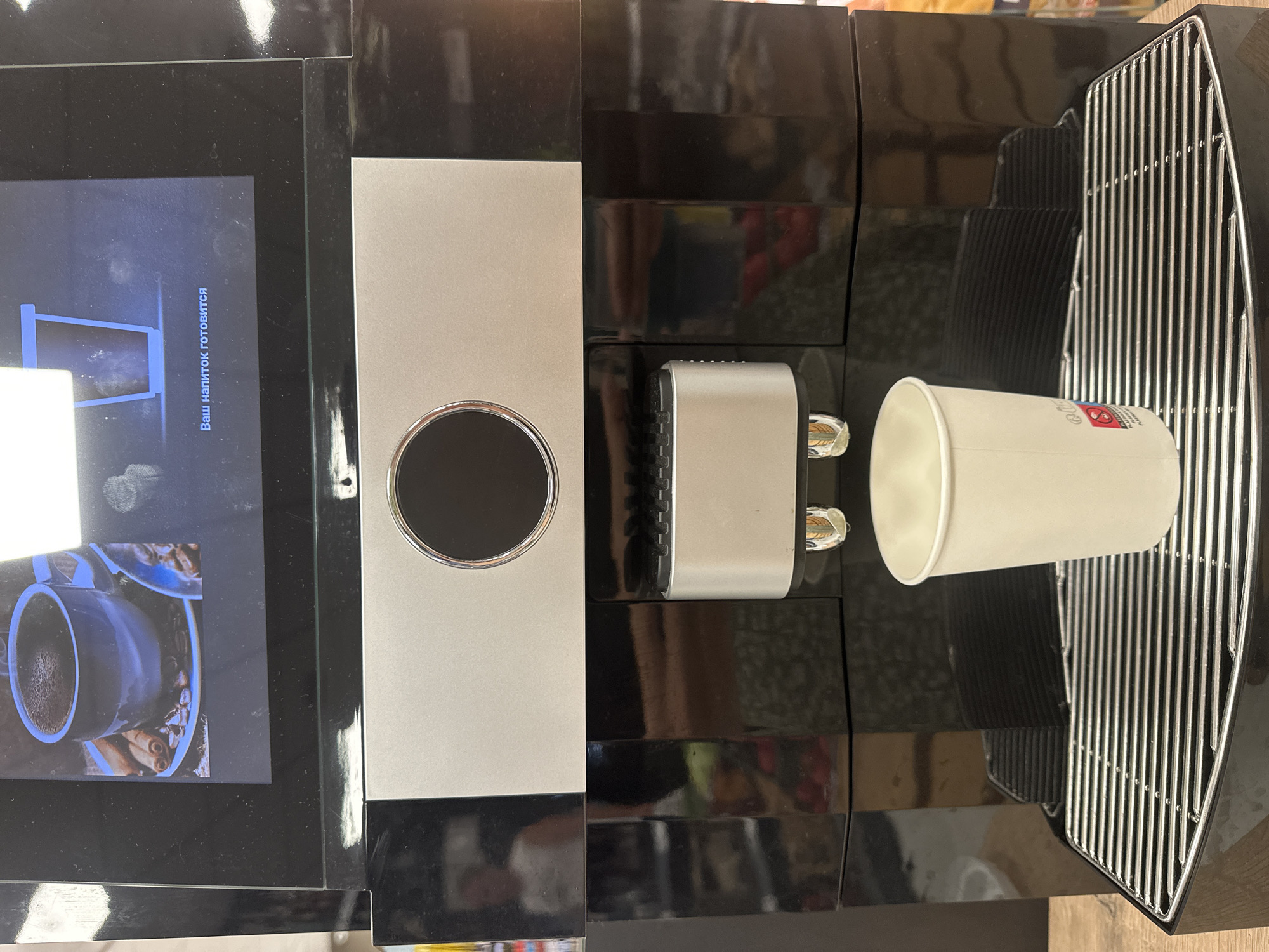 This image shows a close-up, eye-level view of a black automatic coffee machine. The machine has a metallic silver panel on the upper section with a circular black button or sensor in the center. Below this, there is a dispensing spout with two small chrome nozzles. A white paper cup is positioned directly under the spout on a shiny metal drip tray. The screen above the silver panel displays an image of a blue mug of coffee with cinnamon sticks and coffee beans, and text that reads "Ваш напиток готовится" (Your drink is being prepared) in Russian. The background is blurry and appears to be a retail environment with shelves, possibly a supermarket. There is no visible indication of the city of Donji Pobori, Montenegro.