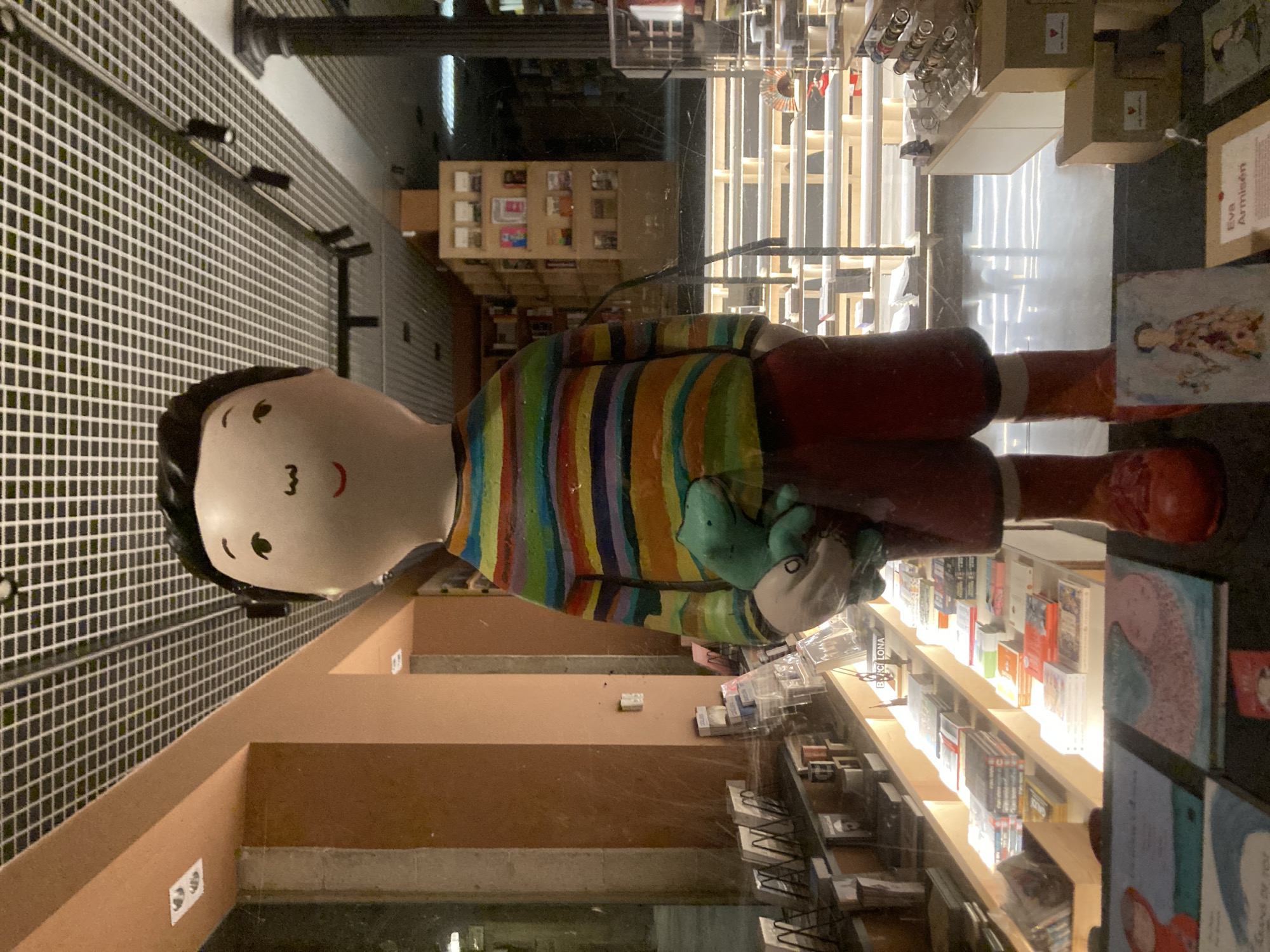 The image depicts the interior of a shop, likely a bookstore, in Barcelona, Spain. A large, doll-like figure of a boy in a colorful striped shirt and red trousers is the central element. The boy is holding a green object, possibly a stuffed animal. Bookshelves filled with books line the interior, indicating the location as a bookstore. Shelves of small objects are visible in the background, adding to the impression of a shop. The grid pattern on the wall suggests the structure is modern.
