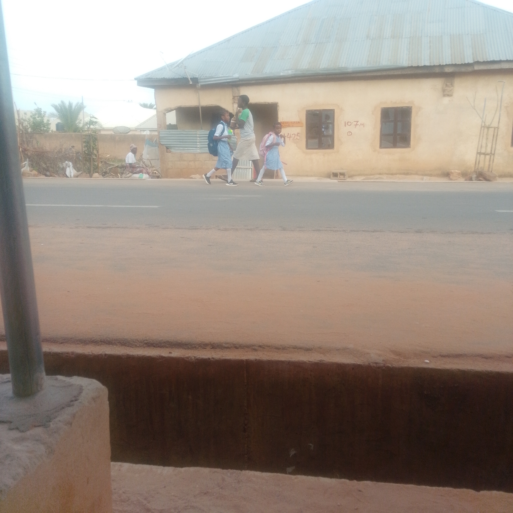 In Gombe, Nigeria, along a paved road, three individuals are walking. Two of them, appearing to be young girls, are wearing matching blue and white checkered dresses with white socks and dark shoes. They both have backpacks; one is navy blue, and the other is pink. A third person, an adult with dark skin and wearing a green shirt and a light-colored skirt, walks between them, facing away from the viewer. They are walking on the right side of the road. In the background, a single-story building with a corrugated metal roof stands. The building is a light tan color and has two dark-paned windows and some graffiti scribbled on its exterior. The ground in front of the building appears to be made of compacted dirt and pebbles, and some vegetation is visible on the left side of the scene. A metal pole is visible on the far left of the image, in the foreground, suggesting it is a street light or utility pole.