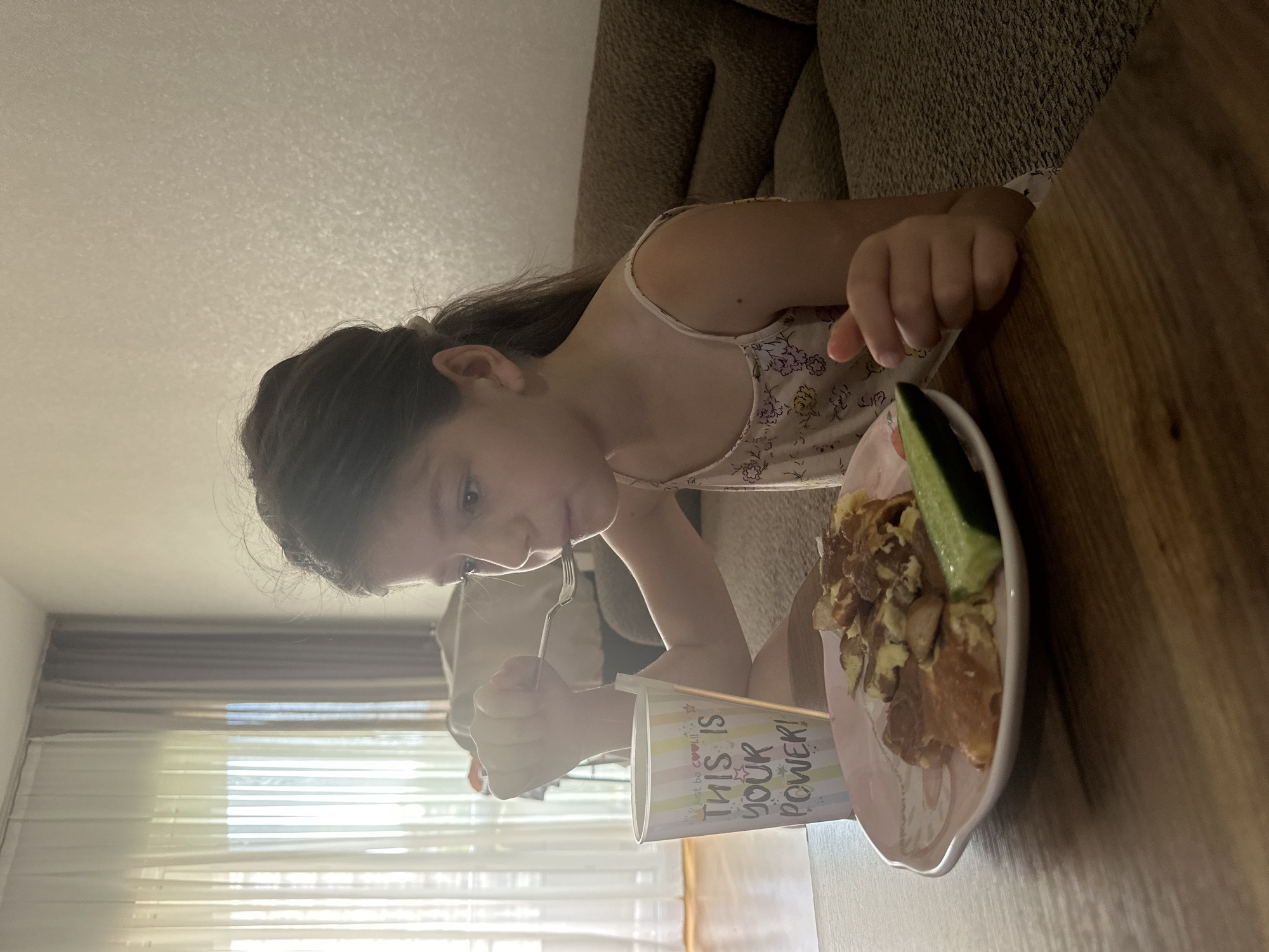 A young girl with brown hair pulled back in a ponytail is seated at a wooden table, eating breakfast. She is wearing a light-colored sleeveless top with a floral pattern. In front of her is a pink plate with scrambled eggs and a slice of cucumber. To her left is a white cup with colorful lettering that reads, "Just be COOL! THIS IS YOUR POWER!". The girl is holding a fork to her mouth, actively eating. Behind her, a brown couch is visible, and to the left, sheer curtains cover a window, allowing natural light to enter the room. The setting appears to be an indoor residential environment, consistent with a home in Esenyurt, Turkey.