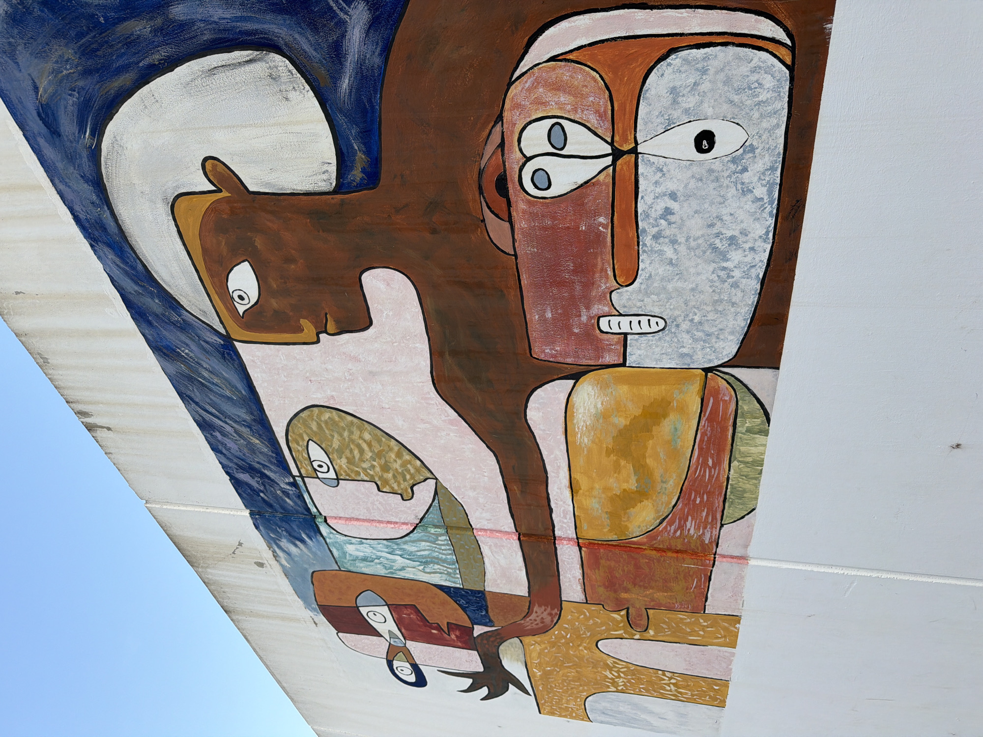 This is a close-up, low-angle shot of a colorful mural painted on a sloped surface, possibly a bridge or overpass, against a clear blue sky. The artwork features abstract figures with stylized faces and bodies, rendered in bold outlines and vibrant colors like brown, red, yellow, and blue. The style suggests a modernist or cubist influence.
In the upper left, a brown, horse-like head with large eyes is depicted within a swirling blue and white background. To its right, a large figure with a reddish-brown face and a textured light blue area for its eye dominates the composition. This figure has small, dark eyes and a mouth with white, linear teeth. Other figures and shapes, rendered in pink, yellow, and blue, are layered and intertwined, creating a sense of depth and complexity. One figure appears to be holding or interacting with another, smaller figure that has wide, dark eyes.
The mural appears to be outdoors, indicated by the clear sky and the way the light falls on the painted surface. The condition of the painted surface suggests it is exposed to the elements, with some signs of wear and tear. The context of Benimàmet-beniferri, Spain, implies an urban or semi-urban setting. The overall mood of the artwork is abstract and somewhat enigmatic, with the stylized figures evoking a sense of narrative or symbolic meaning that is open to interpretation. There is no visible text in the image.