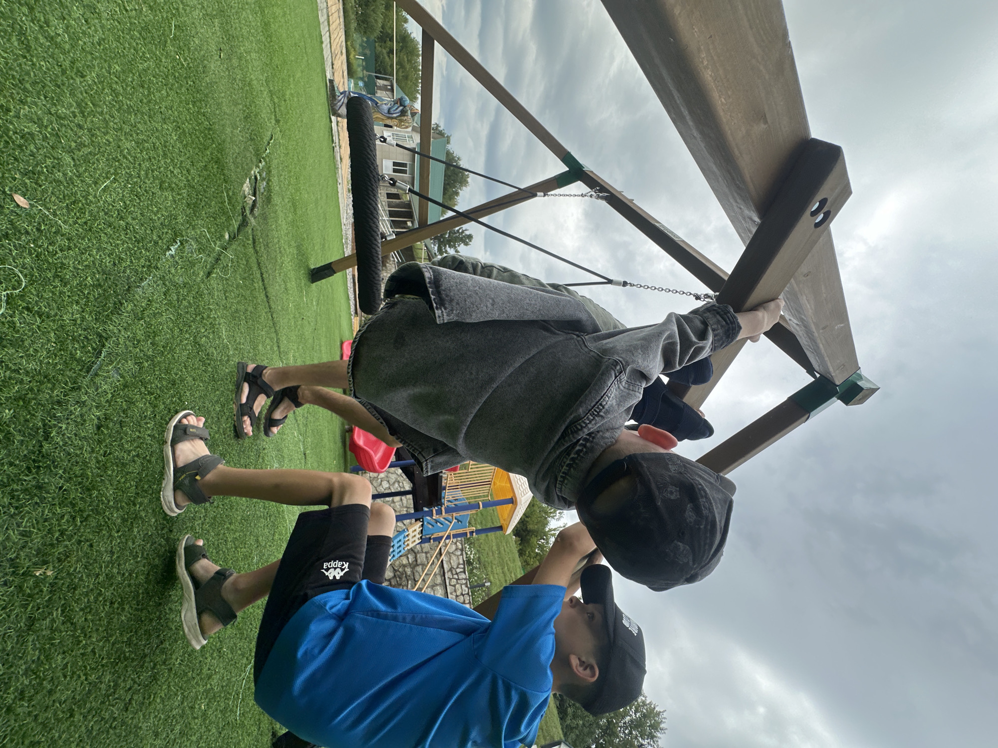 Two boys are playing on a swing set in a yard. The boy on the right is wearing a blue t-shirt and black shorts. The boy on the left is wearing a grey jacket and black shorts. They are both wearing sandals. The swing set is made of wood and has a rope seat. The yard is covered in green artificial turf. In the background, there is a playground with a slide and a climbing frame. There are also houses and trees in the background. The sky is cloudy. This scene is located in the village of Danilovka, Altai Krai, Russia.