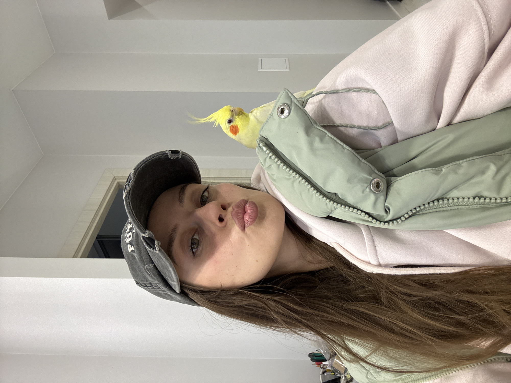A young woman with long brown hair and a gray baseball cap is taking a selfie indoors. She has pursed lips and is wearing a light pink hoodie with a light green puffer jacket over it. A yellow cockatiel with orange cheek patches is perched on the woman's shoulder. The location is likely an apartment in Krasnoyarsk, Russia, given the lighting and interior design, but this cannot be confirmed without further information. The woman is interacting with the camera and her pet bird. The environment appears to be a standard domestic setting.