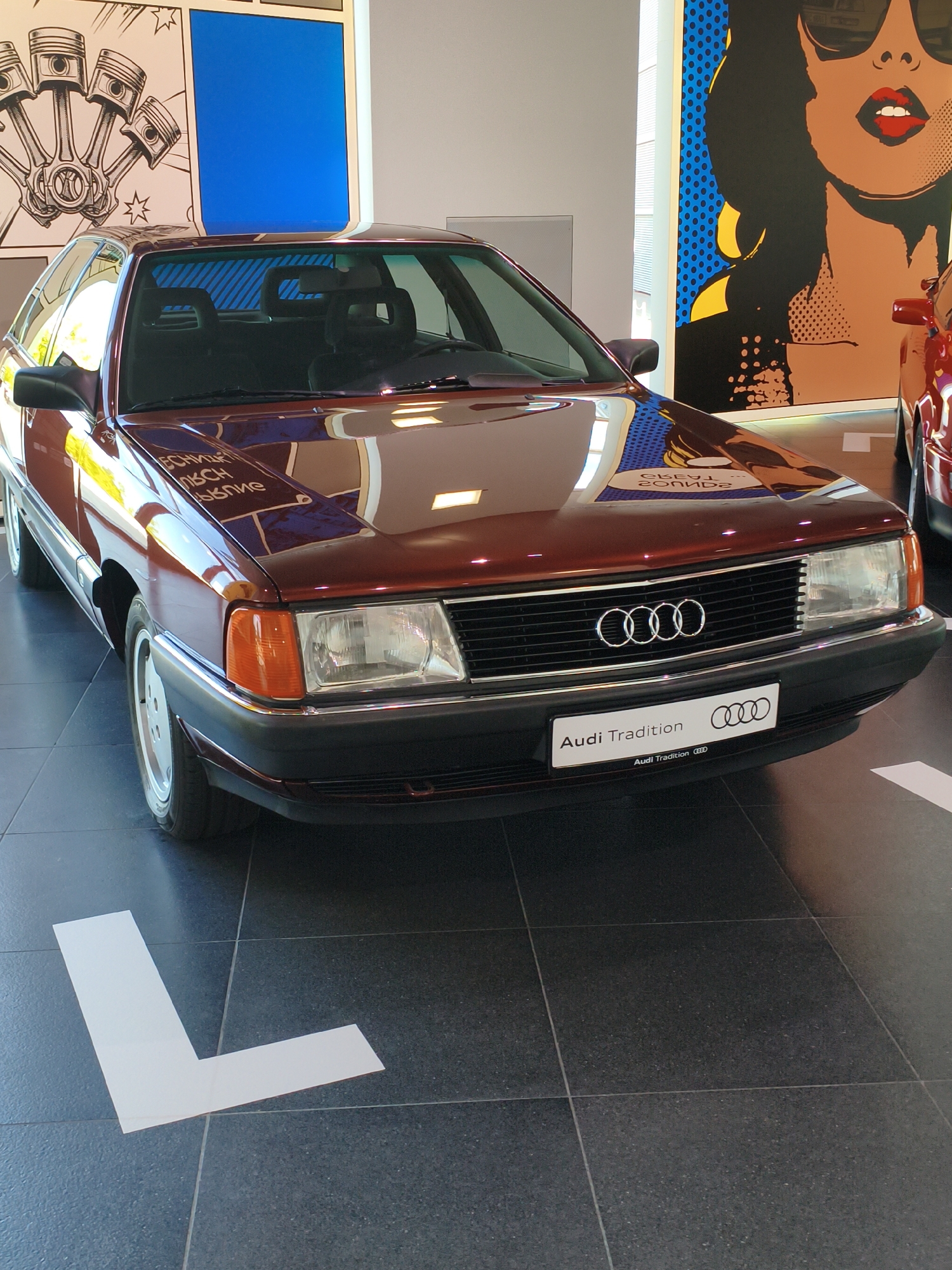The image shows the front of a maroon Audi car, likely a classic model given the "Audi Tradition" badging. The car is parked indoors, on a dark tiled floor with a white "L" shaped marker on the floor. In the background, there are large, colorful graphic art pieces on the walls, one depicting engine pistons and another a stylized portrait of a woman. The lighting suggests an indoor showroom or exhibition space. The car's hood reflects the surrounding environment, including parts of the wall art and lighting fixtures. The presence of an "Audi Tradition" badge indicates this might be a display at the Audi Forum in Ingolstadt, Germany, which houses a museum dedicated to Audi's history. There are no people visible in the image.