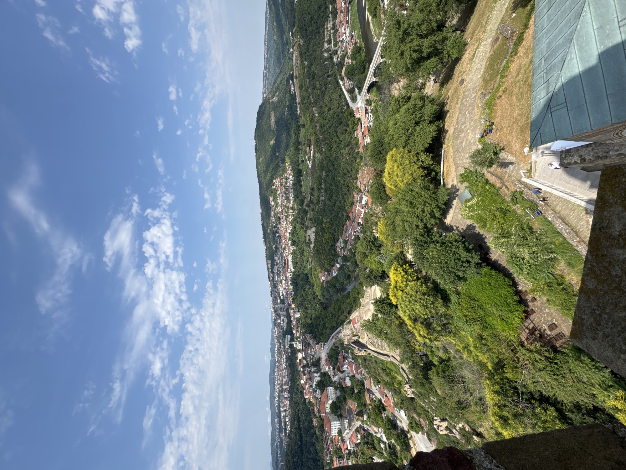 This image provides a panoramic view of Veliko Tarnovo, Bulgaria, taken from a high vantage point, likely within or near Tsarevets Fortress. The scene captures the city nestled within a valley, characterized by rolling hills covered in lush green trees. Red-tiled roofs of numerous buildings are visible, dotting the landscape and clustered in dense residential areas. A prominent river, likely the Yantra River, meanders through the valley, crossed by a white arched bridge in the mid-ground. The foreground features stone structures and vegetation, suggesting the viewpoint is from a historic fortification or a similar elevated tourist spot. The sky is a bright blue with scattered white clouds, indicating a clear day. Several people are visible on pathways and in areas around the historical structures in the foreground, suggesting tourism activity. The overall impression is one of a historic European city surrounded by natural beauty.