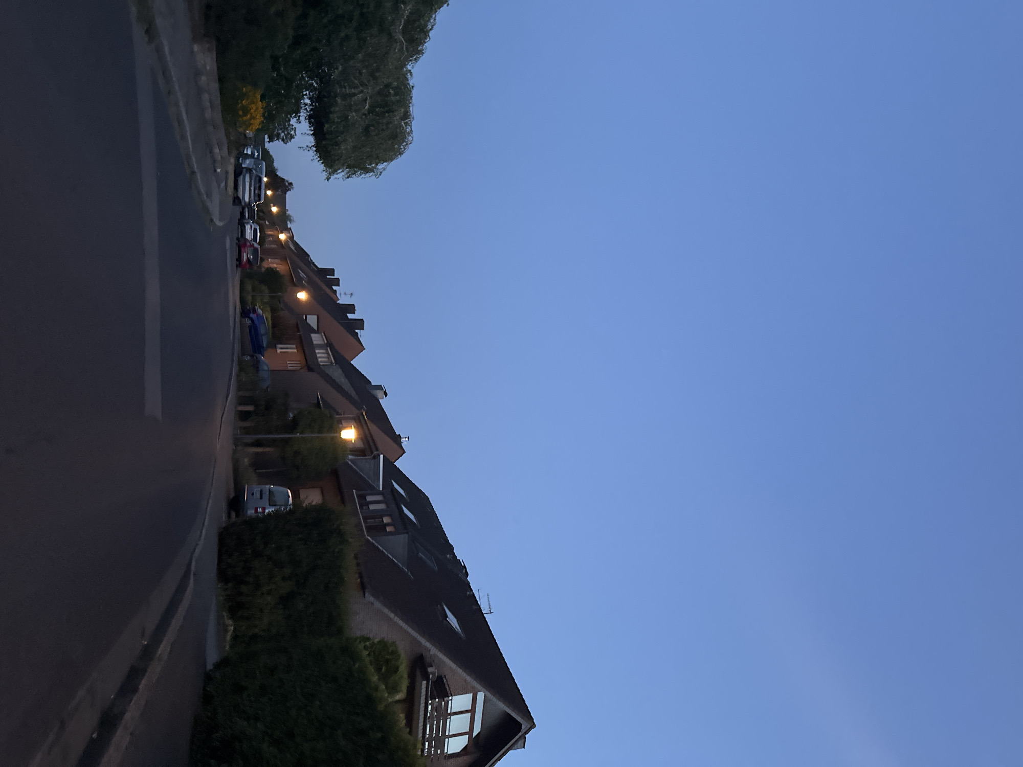 The image depicts a quiet residential street in Krefeld, Germany, at dusk. A row of traditional German houses with pitched, dark roofs lines the right side of the street, some with visible dormer windows and chimneys. Streetlights illuminate the scene, casting a warm glow on the pavement and the parked cars. Several vehicles, including a silver van, a red hatchback, and a blue sedan, are parked along the side of the road. On the left side of the street, lush green trees and hedges create a natural barrier. The sky above is a clear, deep blue, indicating the transition from daylight to night. The overall impression is one of tranquility and suburban life in Krefeld.