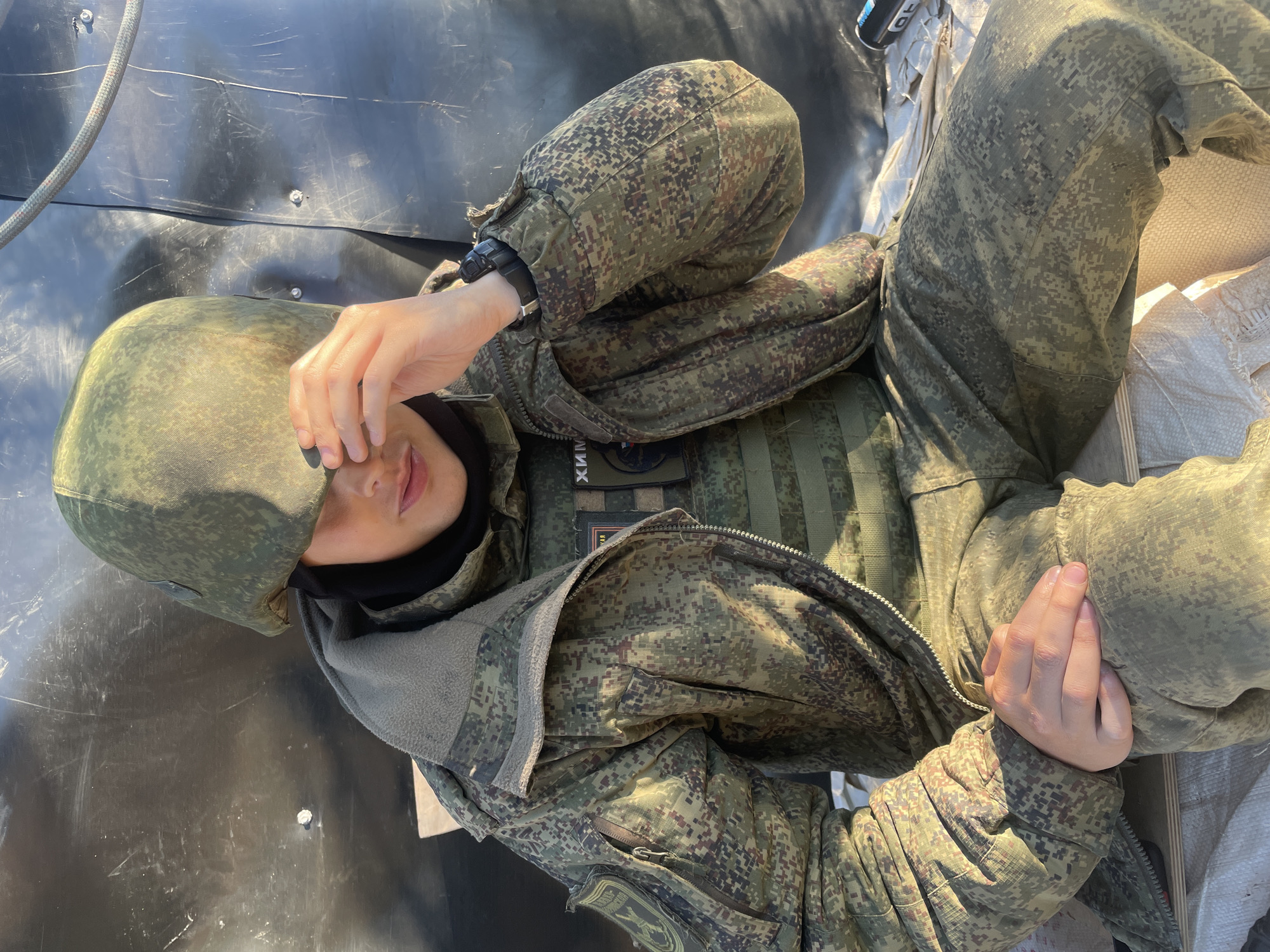 A person wearing a camouflage military uniform and helmet sits on what appears to be a wooden surface covered with white fabric. The person is looking down and has their right hand covering their eyes. The background consists of a dark, textured surface with some metallic elements and a rope. Patches are visible on the uniform, including one with Cyrillic text. The lighting suggests an outdoor setting with sunlight casting shadows. The person's left hand rests on their lap.