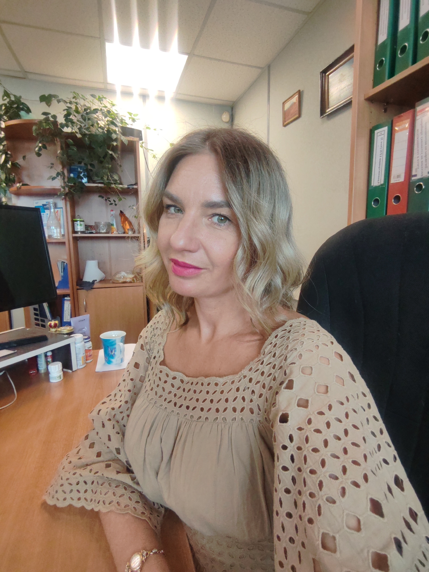 The image shows a middle-aged woman sitting at a desk in what appears to be an office. The woman has blonde, wavy hair, green eyes, and is wearing bright pink lipstick. She is wearing a beige, long-sleeved blouse with decorative cutouts. A gold wristwatch is visible on her left wrist. She looks directly at the camera.
The office setting includes a wooden desk cluttered with office supplies, including a computer monitor, a cup, and several small bottles. A bookshelf filled with plants and various objects is visible behind the desk, and there is a cabinet stocked with binders to the right. The lighting is bright, seemingly from overhead fluorescent lights. Two small, framed pictures hang on the wall in the background. There is a black office chair partially visible on the right of the frame. The city and state are unknown.