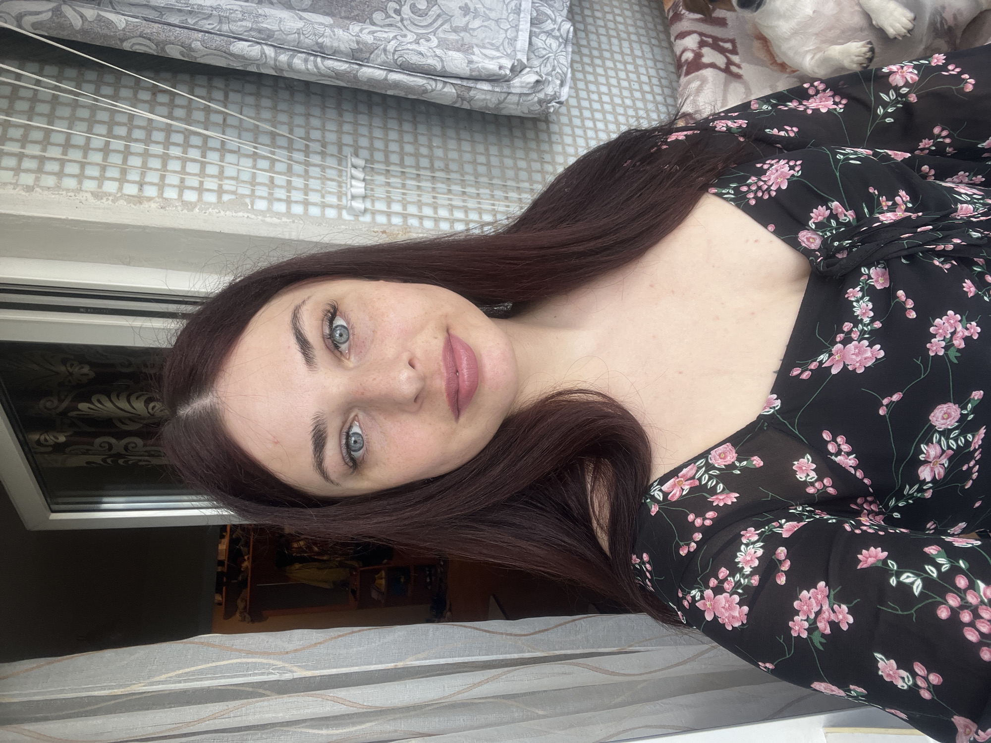 A young woman with long, auburn hair and light blue eyes is pictured in a selfie. She is wearing a black floral dress with pink flowers. Her lips are painted a muted pink. Behind her is a window with patterned curtains and a view of what appears to be a building exterior. The location is indicated as Chaikovsky, Russia. A patterned cushion is also visible in the background. There is a dog in the upper right corner of the image.
