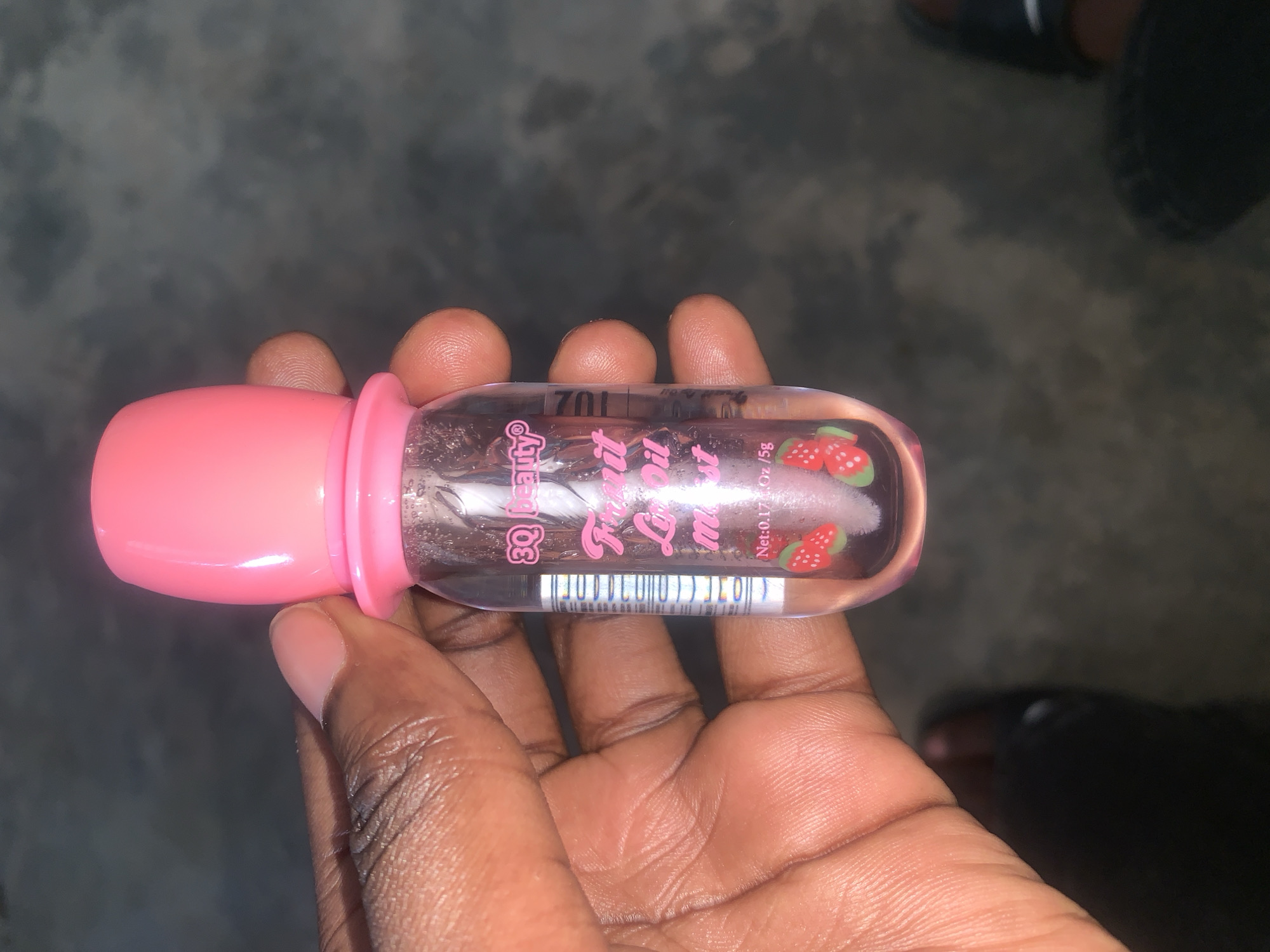 A dark-skinned hand holds a clear bottle with a pink cap and pink accents on the label. The label reads "Beauty Kiss Lip Oil" and displays strawberry images. The bottle contains a transparent liquid with visible particulates. The background is blurry and indistinct, showing a gray surface and a glimpse of black footwear. The location is New Stadium, Nigeria.