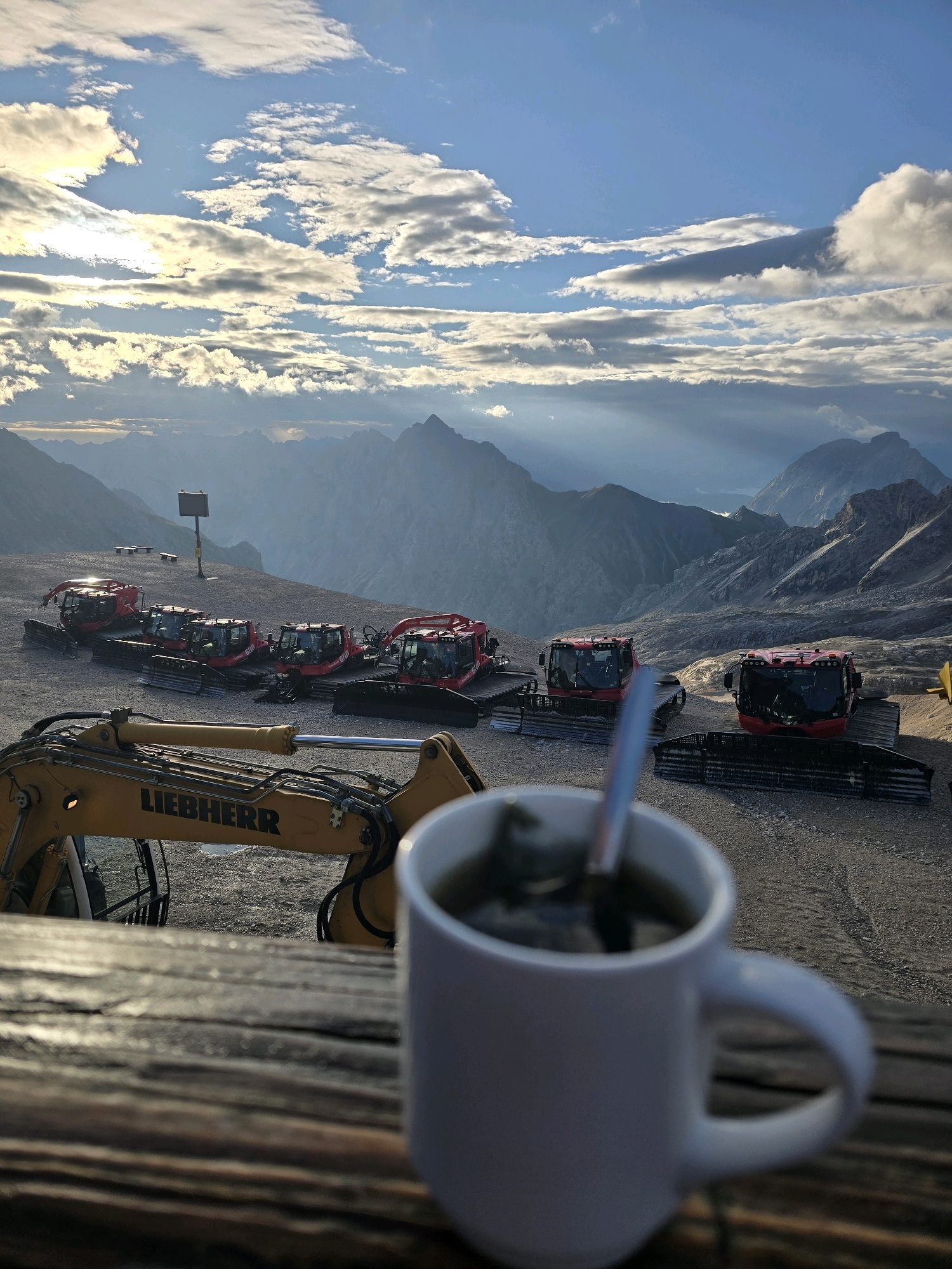 This is a wide-angle, eye-level shot taken in the city of Garmisch-Partenkirchen, Germany. The image features a panoramic view of a mountainous landscape under a partly cloudy sky. In the foreground, a white mug with a spoon in it is placed on a wooden surface. In the middle ground, several red snow groomers are parked on a gravel surface, with a yellow Liebherr excavator to the left. The background consists of rugged mountain ranges receding into the distance, with sunlight breaking through the clouds in some areas.
